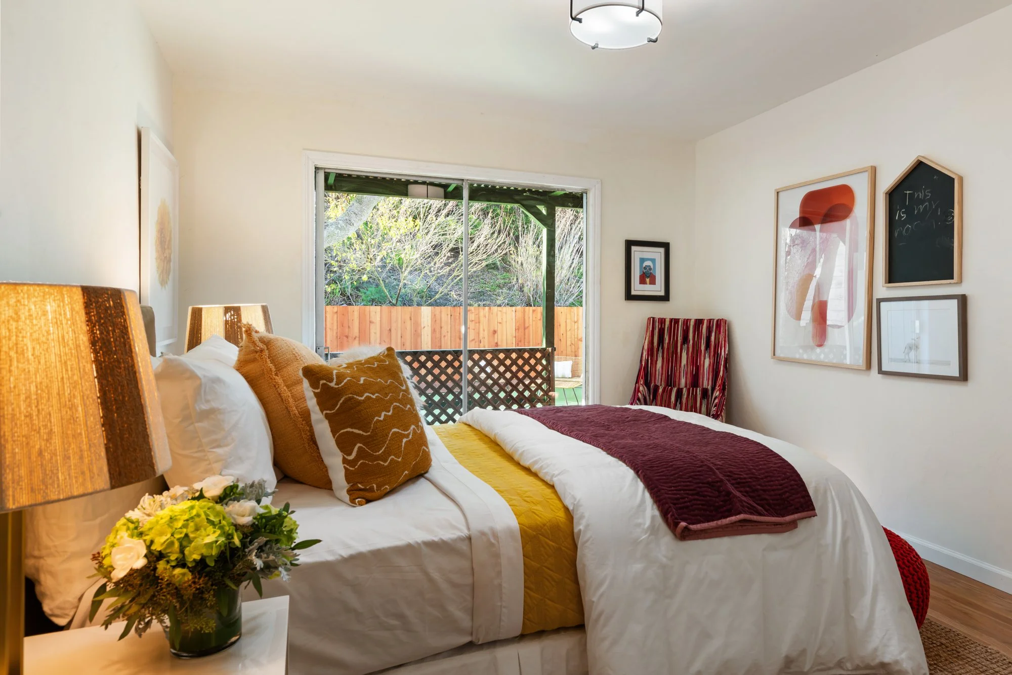 Cozy bedroom with a large bed, mustard and brown pillows, a window with a view of a fenced backyard and trees, and wall art.
