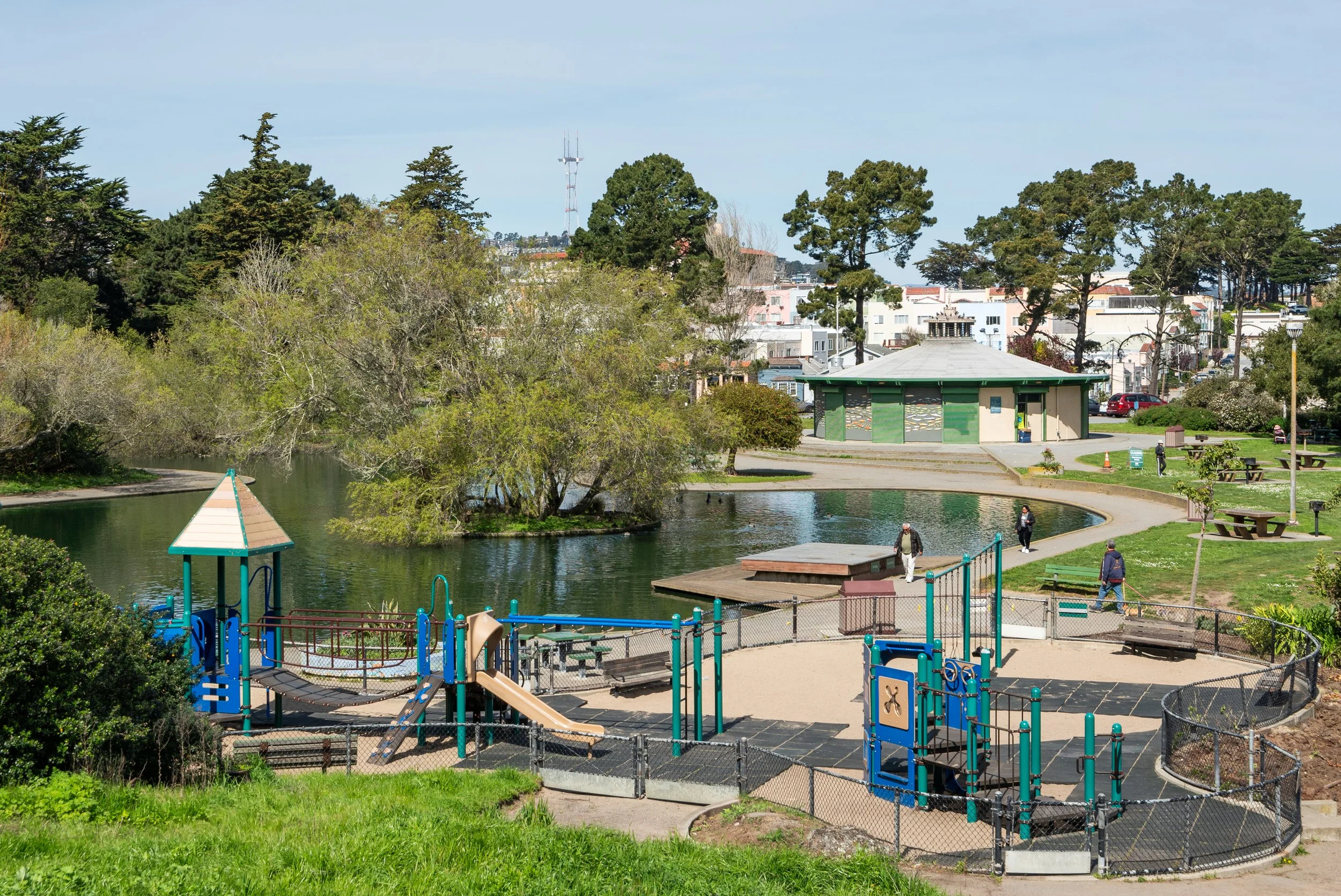 A park with a playground next to a small lake, surrounded by trees and grassy areas. There are a few people walking along the paved paths, and a building with green doors in the background. The sky is clear with some clouds.