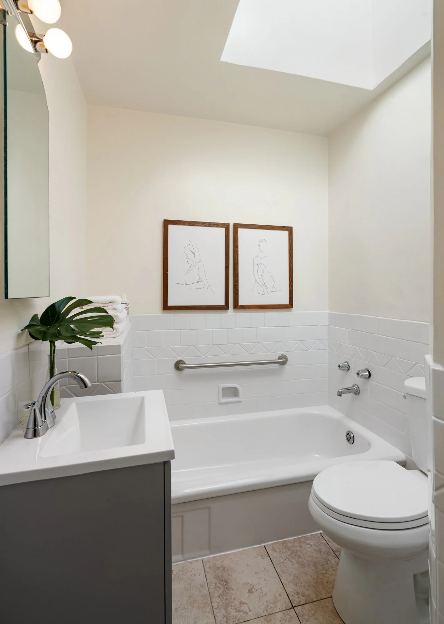 Bathroom with a bathtub, a toilet, a sink with a plant on the counter, two framed line drawings on the wall, a mirror, and a skylight overhead.