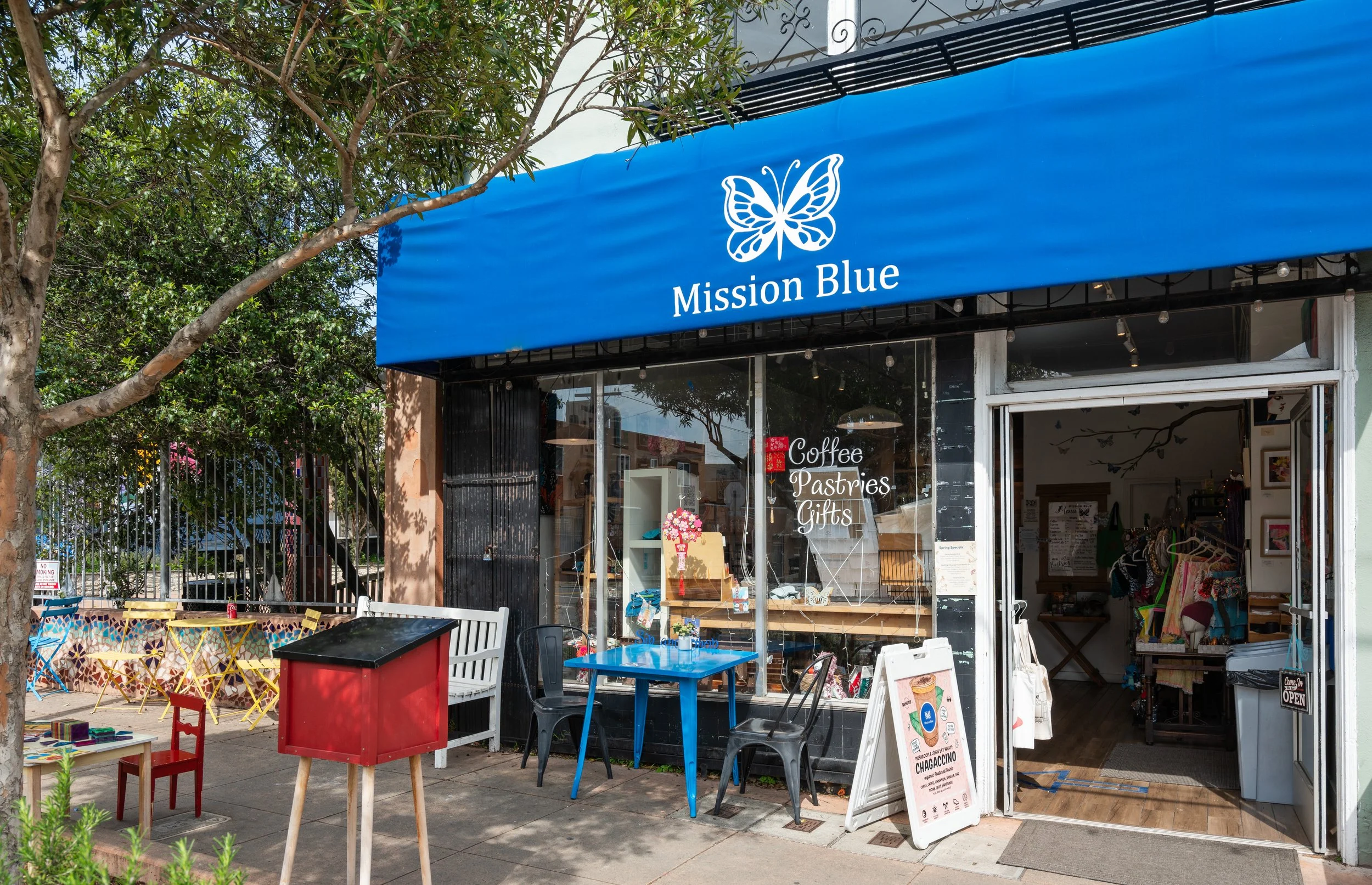 Exterior of a shop called Mission Blue with a blue awning and a butterfly logo. The shop offers coffee, pastries, and gifts. There are outdoor tables and chairs, and the shop's entrance is open, revealing inside merchandise.