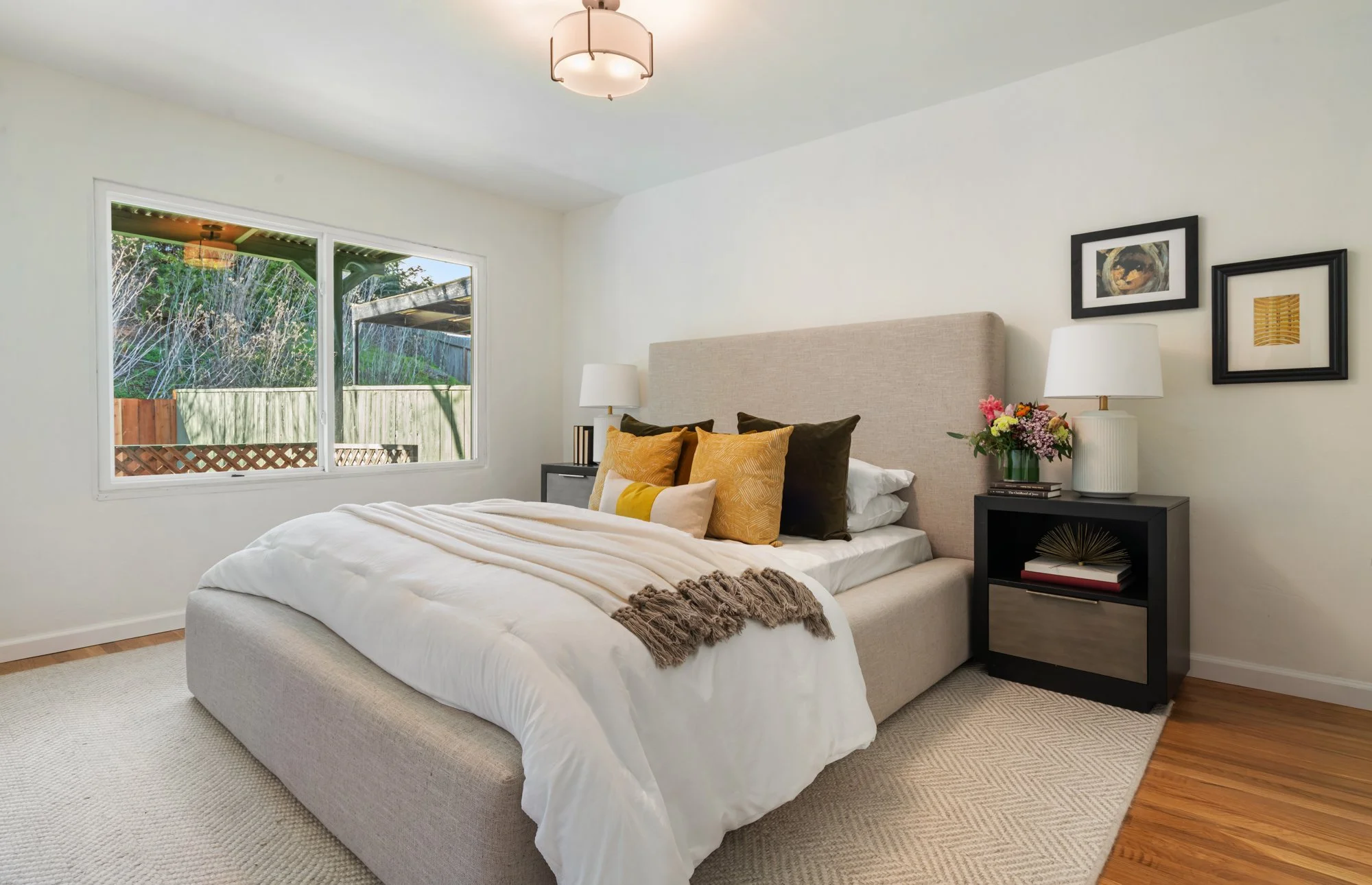 A neatly made bed with a beige upholstered headboard, layered with white and gold pillows, a blanket, and a folded throw, flanked by black nightstands with lamps and a flower arrangement, next to a window with a view of a backyard.