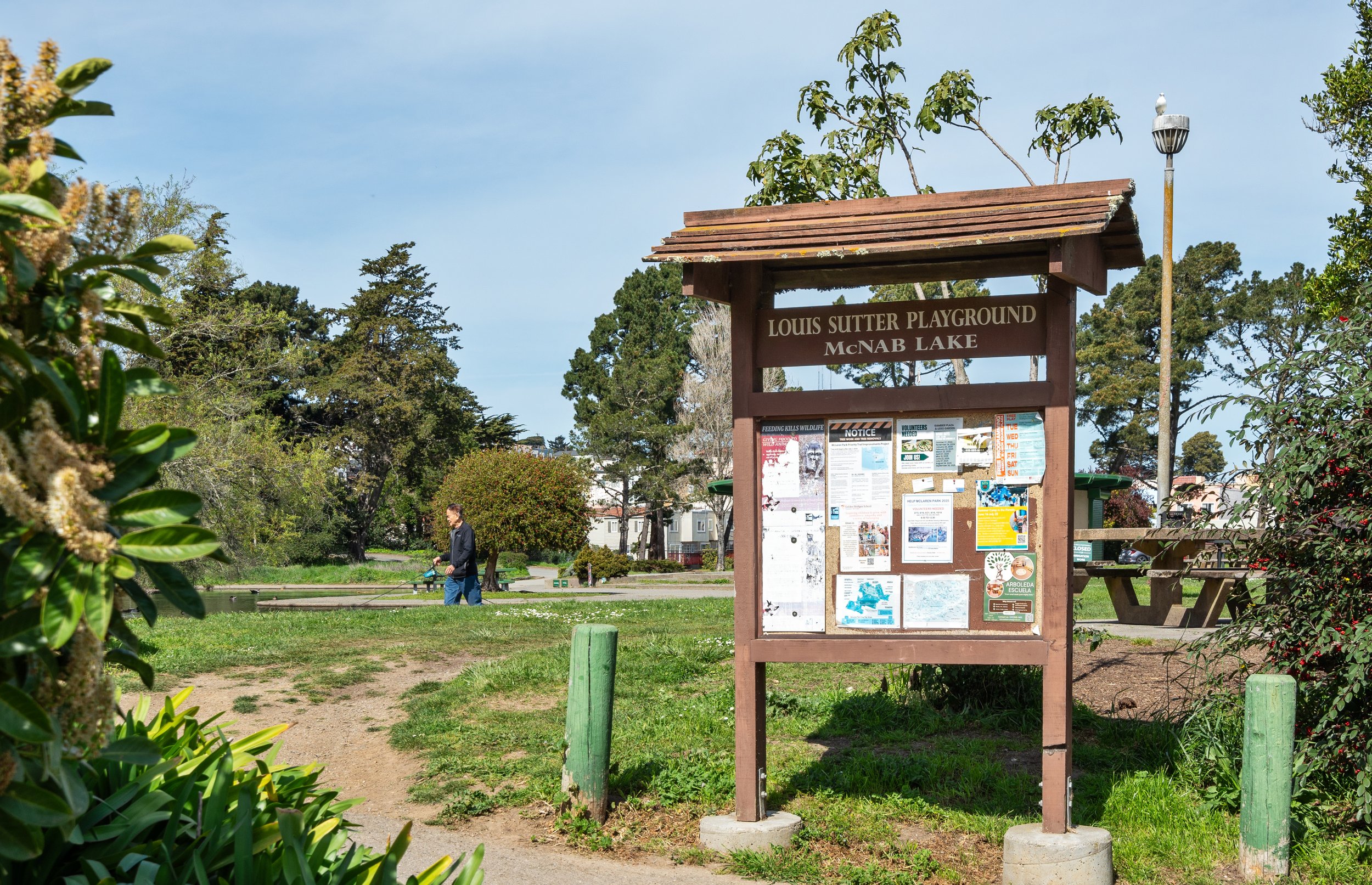 A park with a wooden signboard that reads 'Louis Sutter Playground McNAB Lake.' The sign has notices and flyers attached. There are trees, bushes, and a person walking near the playground in the background. The sky is partly cloudy.
