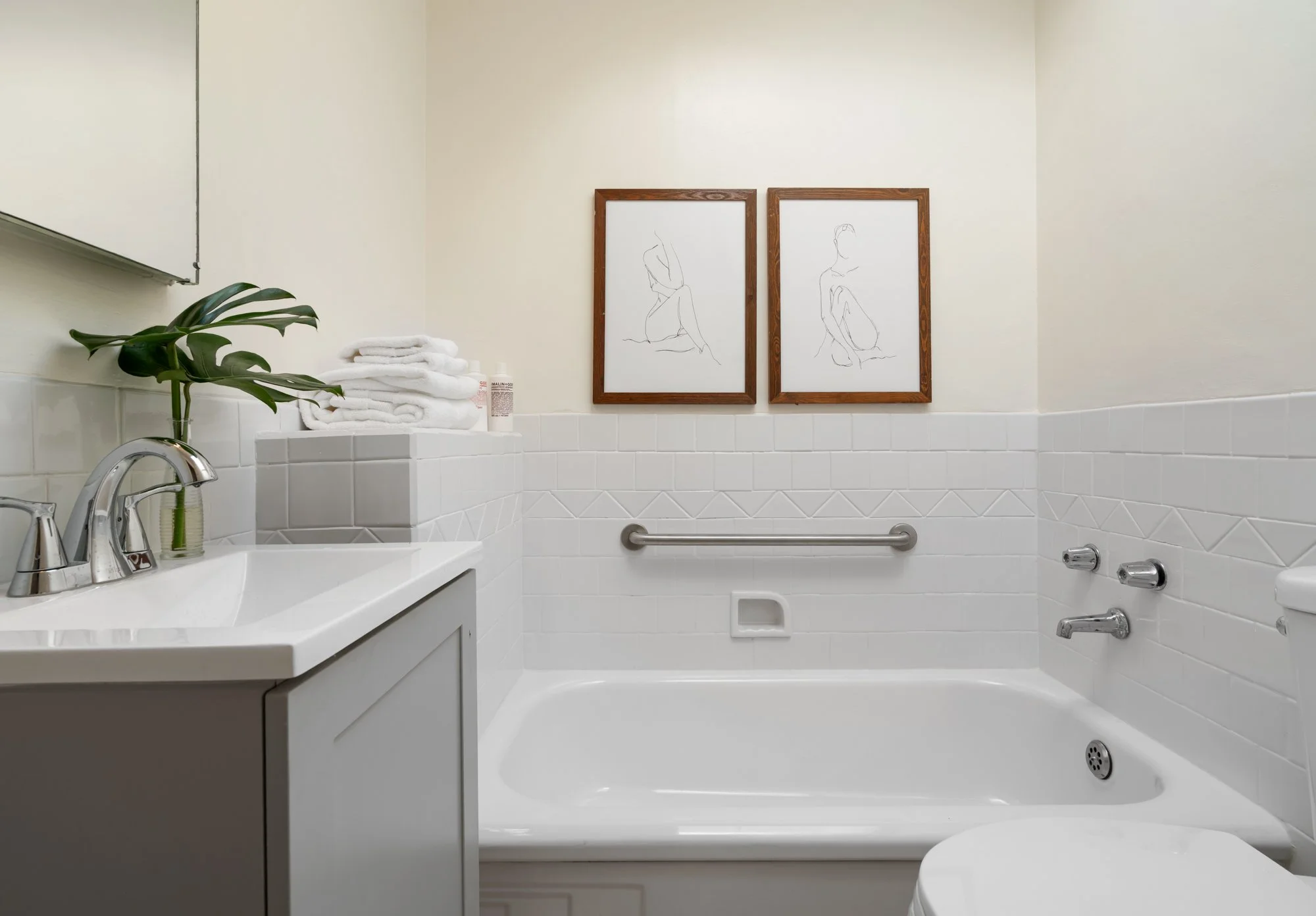 Bathroom with white tiled walls, a bathtub with grab bar, a sink with a faucet, a potted plant, folded towels, and framed line art pictures of a woman on the wall.