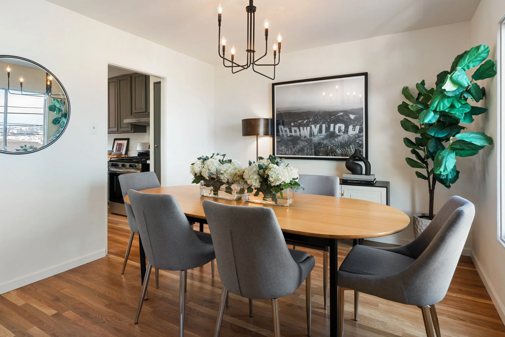 A dining room with a wooden table surrounded by six gray upholstered chairs. A large black and white photograph hangs on the wall, flanked by a tall lamp and a decorative black vase. A green potted plant stands in the corner. A chandelier hangs from 