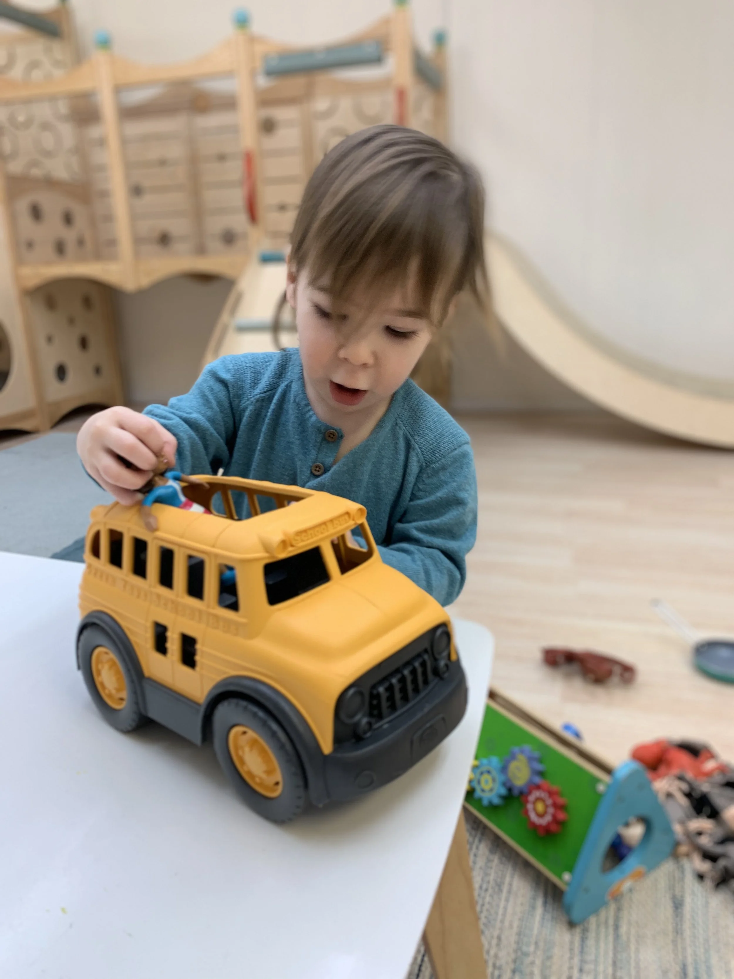 A small child plays with a yellow toy bus in a calm pleasing space filled with wooden toys.