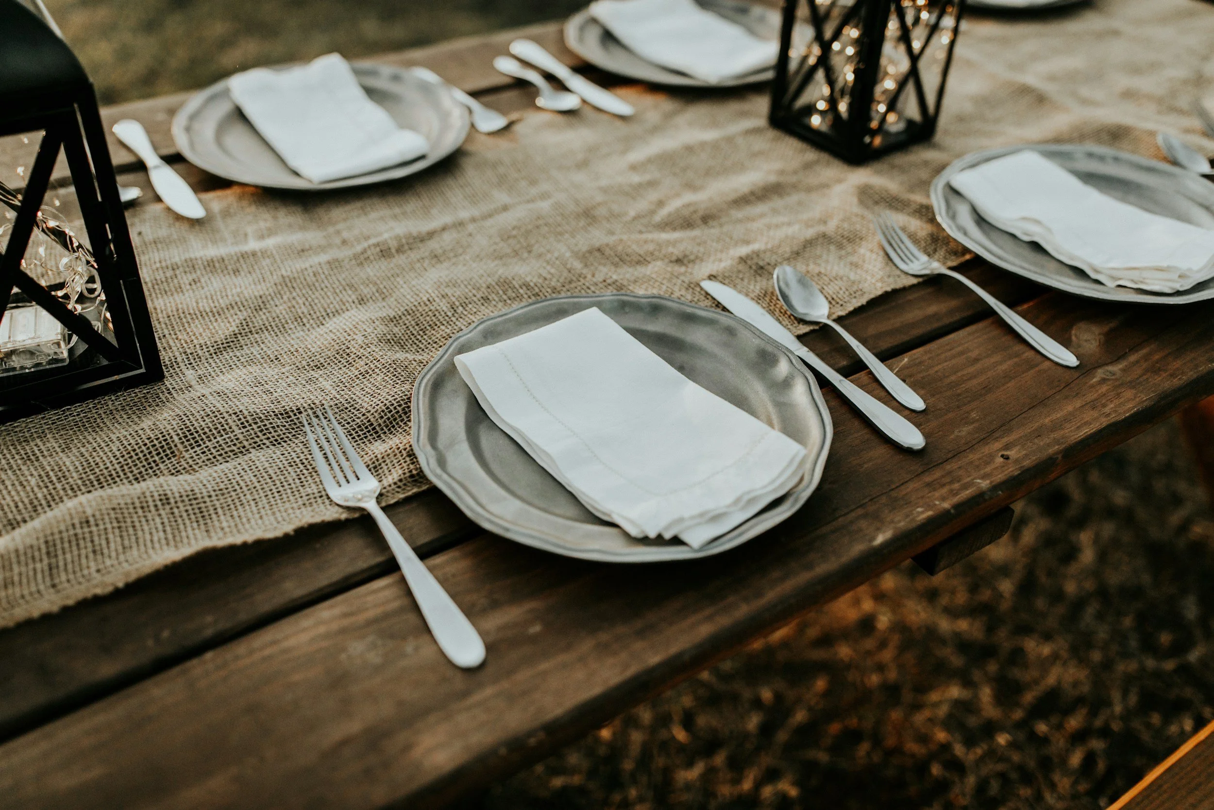 Dining table set outdoors with silver plates, white napkins, and silverware, decorated with a burlap table runner and black lanterns.