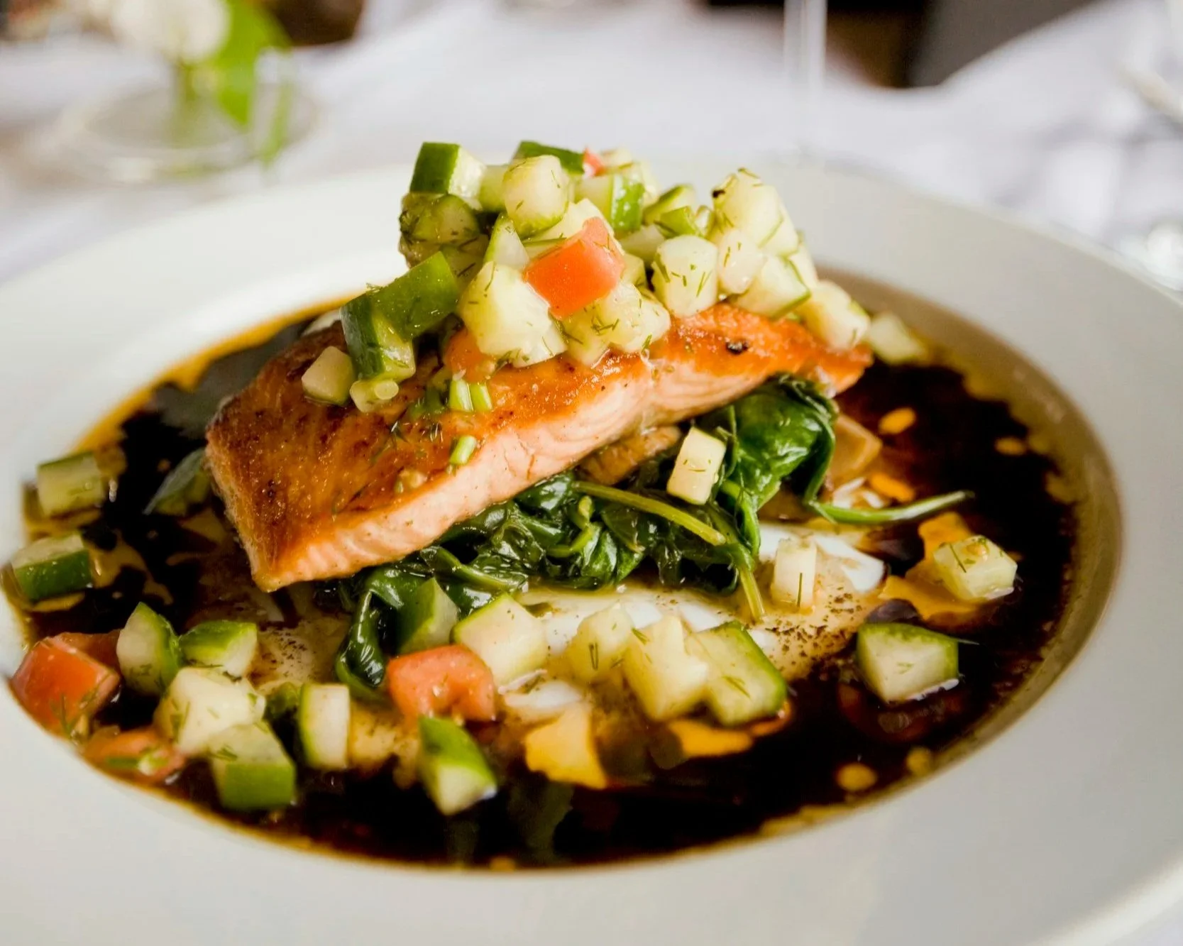 A plated dish with seared salmon topped with a cucumber and carrot relish, served over sautéed greens in a dark sauce.