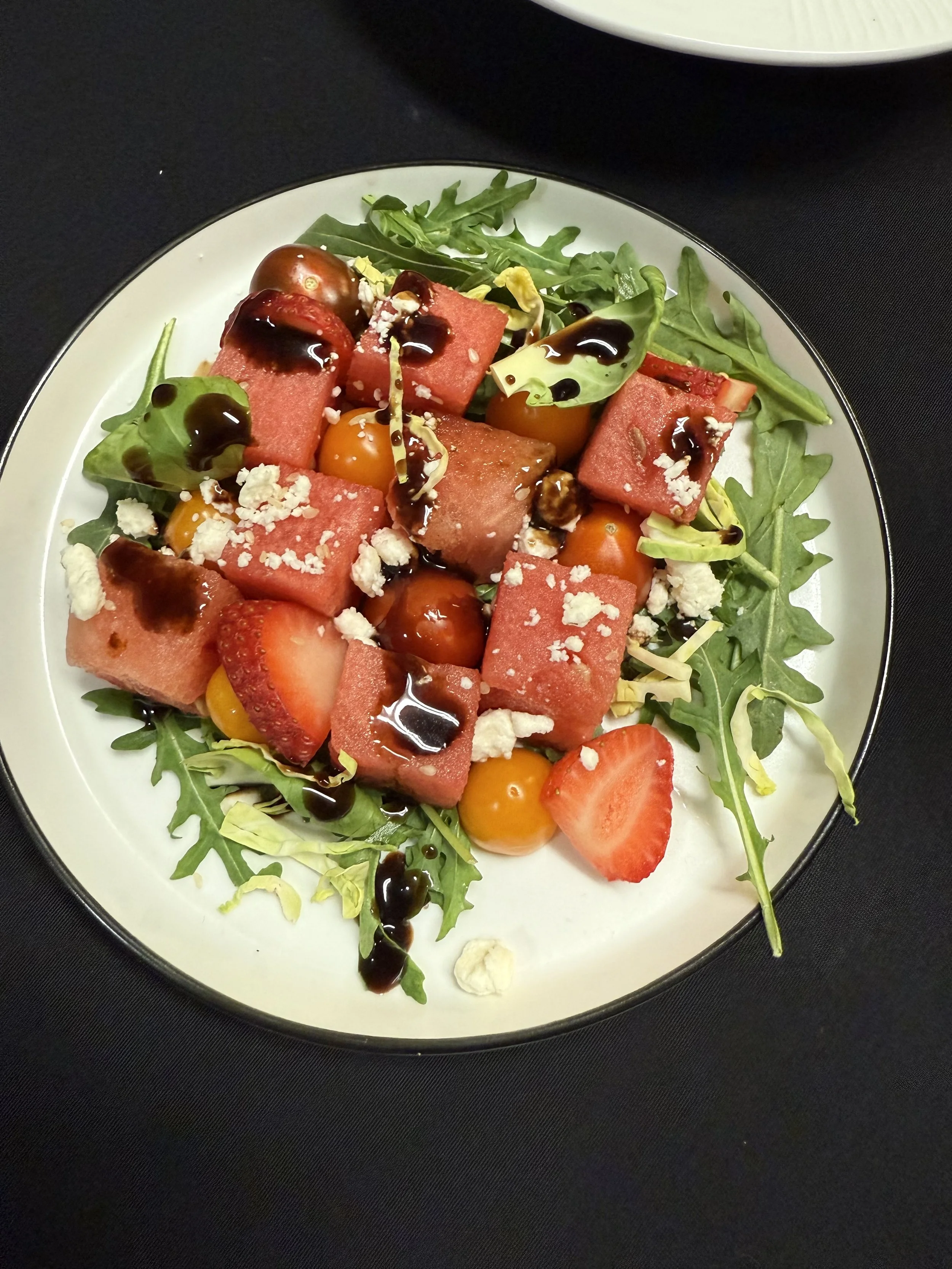 A plate of salad with watermelon cubes, cherry tomatoes, strawberries, arugula, chopped lettuce, crumbled cheese, and a drizzle of balsamic glaze.