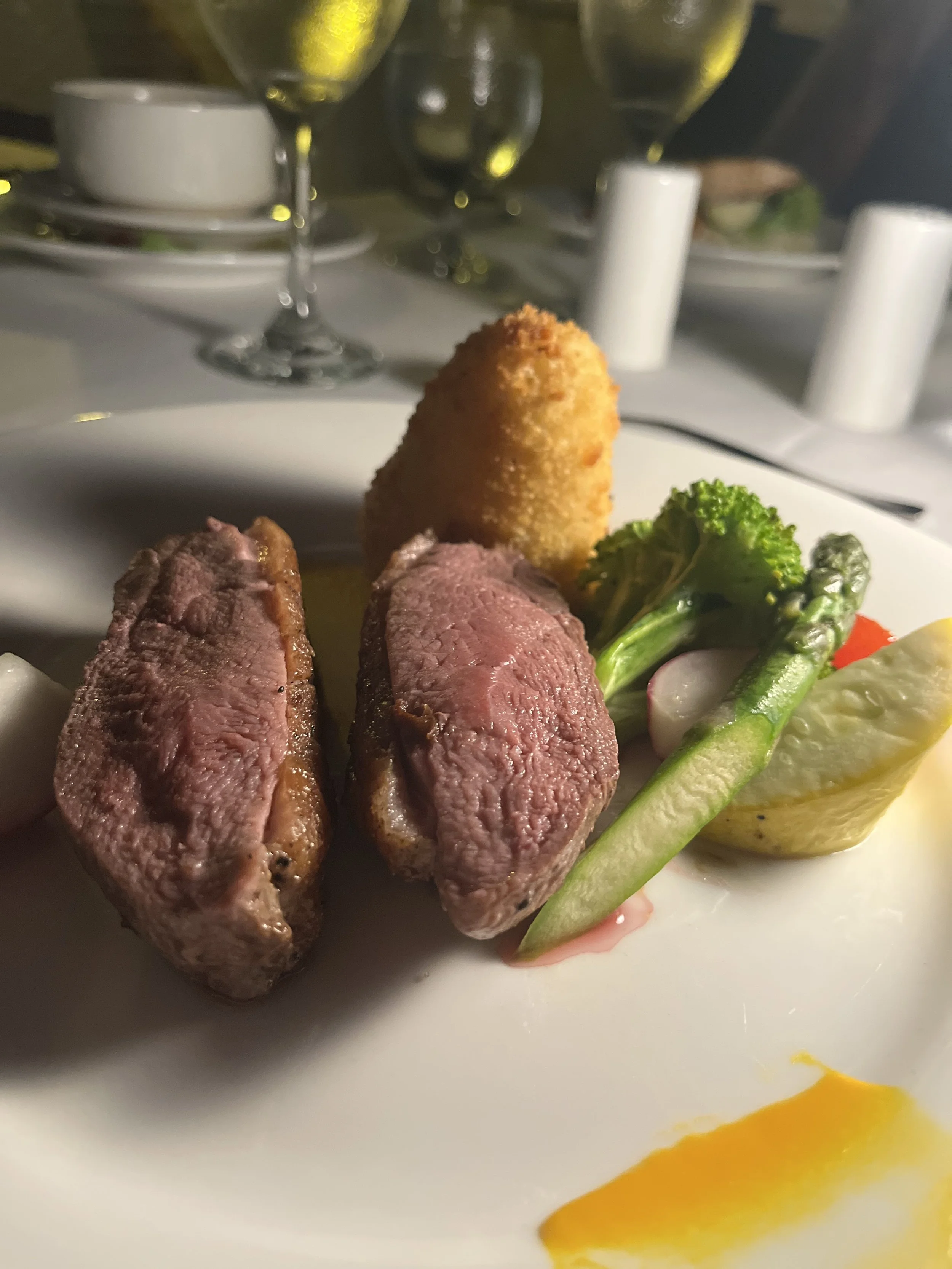 A gourmet dish featuring two slices of medium-rare cooked steak, a breaded croquette, and assorted fresh vegetables including broccoli, radish, snap peas, and lemon wedge, served on a white plate with a yellow sauce accent, in a fine dining setting with glasses and candles in the background.