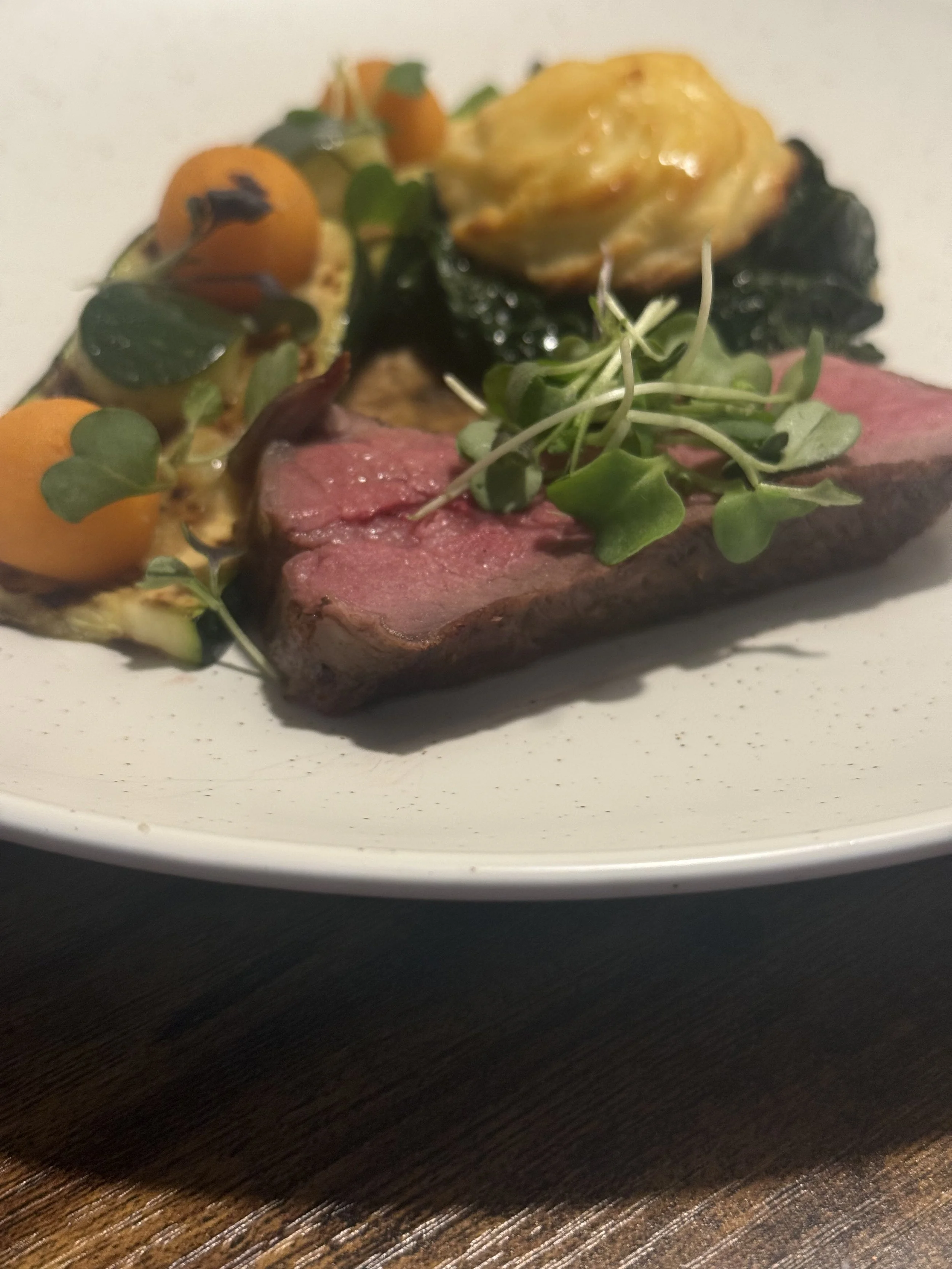 A gourmet dish features a piece of cooked steak topped with microgreens, accompanied by roasted vegetables including cherry tomatoes, zucchini, and a yellow pattypan squash, served on a white plate.