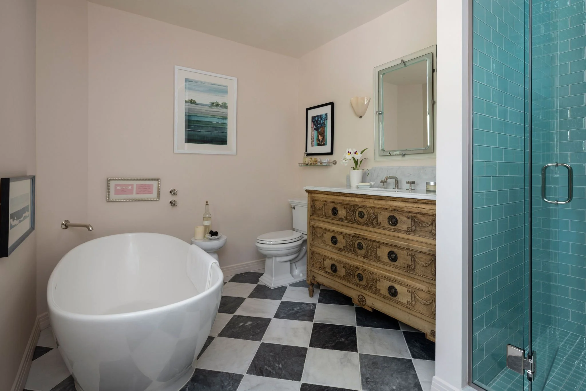 Bathroom featuring a freestanding white bathtub, a toilet, a vintage wooden dresser with a sink, framed artwork, a mirror, a small shelf with decorative items, a potted plant, and a glass shower with teal tiles. Charleston interior designer. 