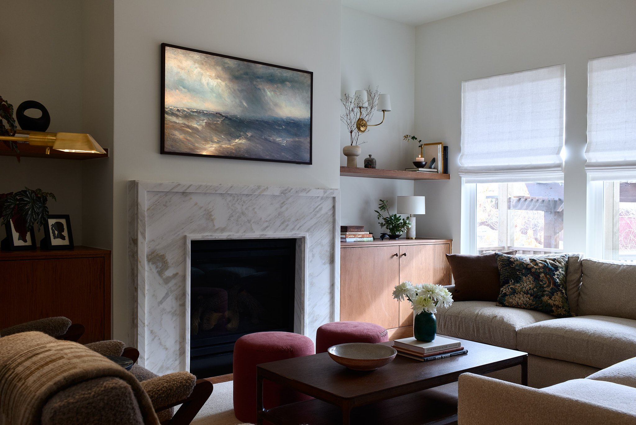 Living room with fireplace, framed painting of ocean waves, beige sofa with patterned pillows, wooden coffee table with books and flowers, window with white shades, wooden sideboard, decorative shelf with vases, lamps, and plants.