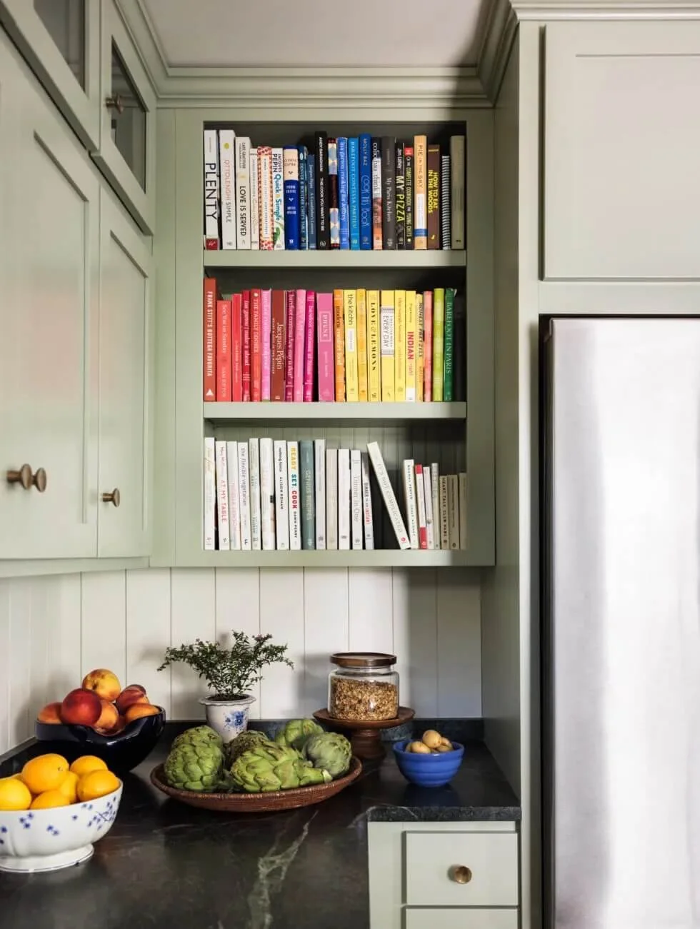 Kitchen corner with green cabinetry, a black countertop, and a refrigerator. Open shelves filled with colorful books and a table with fruits, artichokes, a small potted plant, a jar of grains, and a small blue bowl of onions.