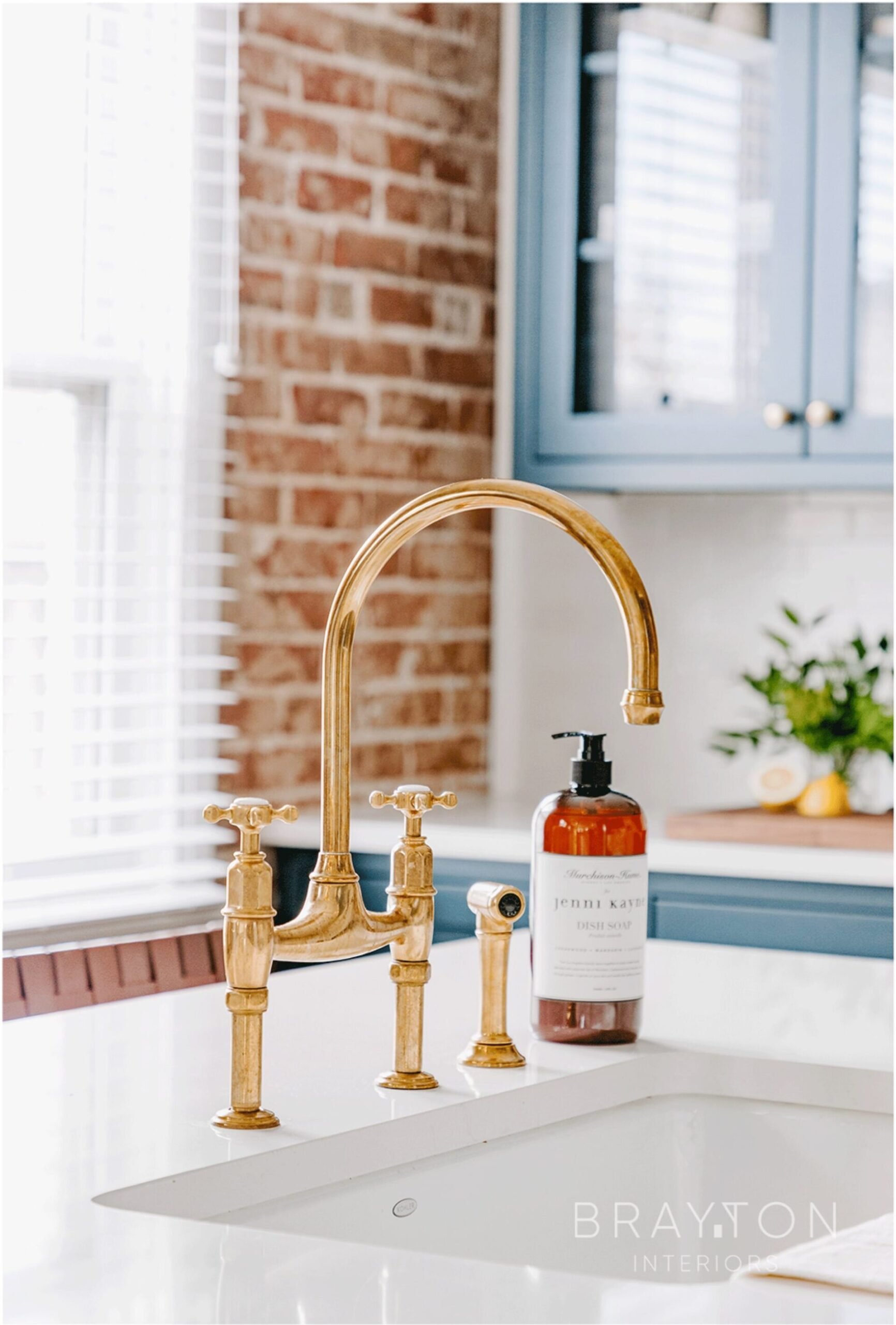 A kitchen sink with a gold faucet with two handles, a soap dispenser, a brick wall, and blue cabinets in the background.