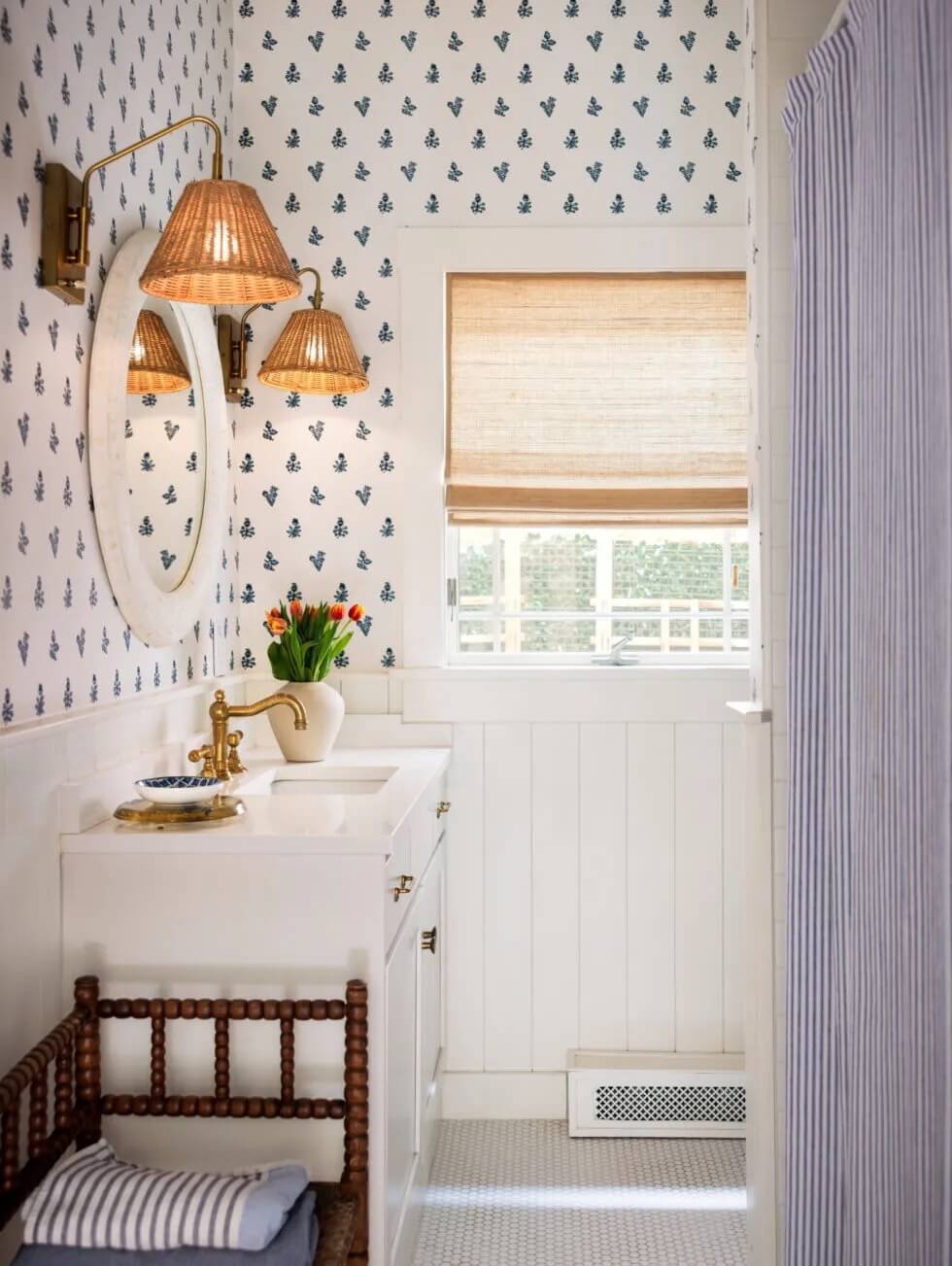 Small bathroom with a vintage style featuring a white vanity with gold fixtures, a mirror, and wicker lampshades. There is a window with a hidden blind, a flower vase with tulips, and a wooden chair with a striped pillow. The walls are decorated with