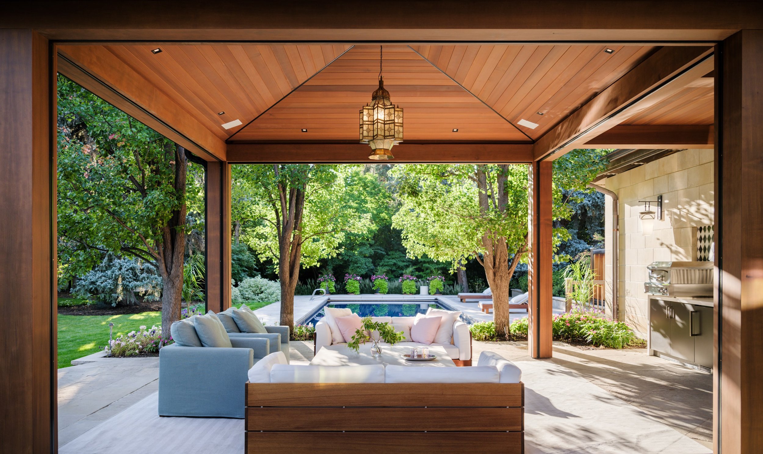 Covered patio with wooden ceiling, outdoor furniture including a white sofa and light blue armchairs, overlooking a garden with trees and a swimming pool in the background. Bonacina outdoor furniture through Charleston Interior Designer. 