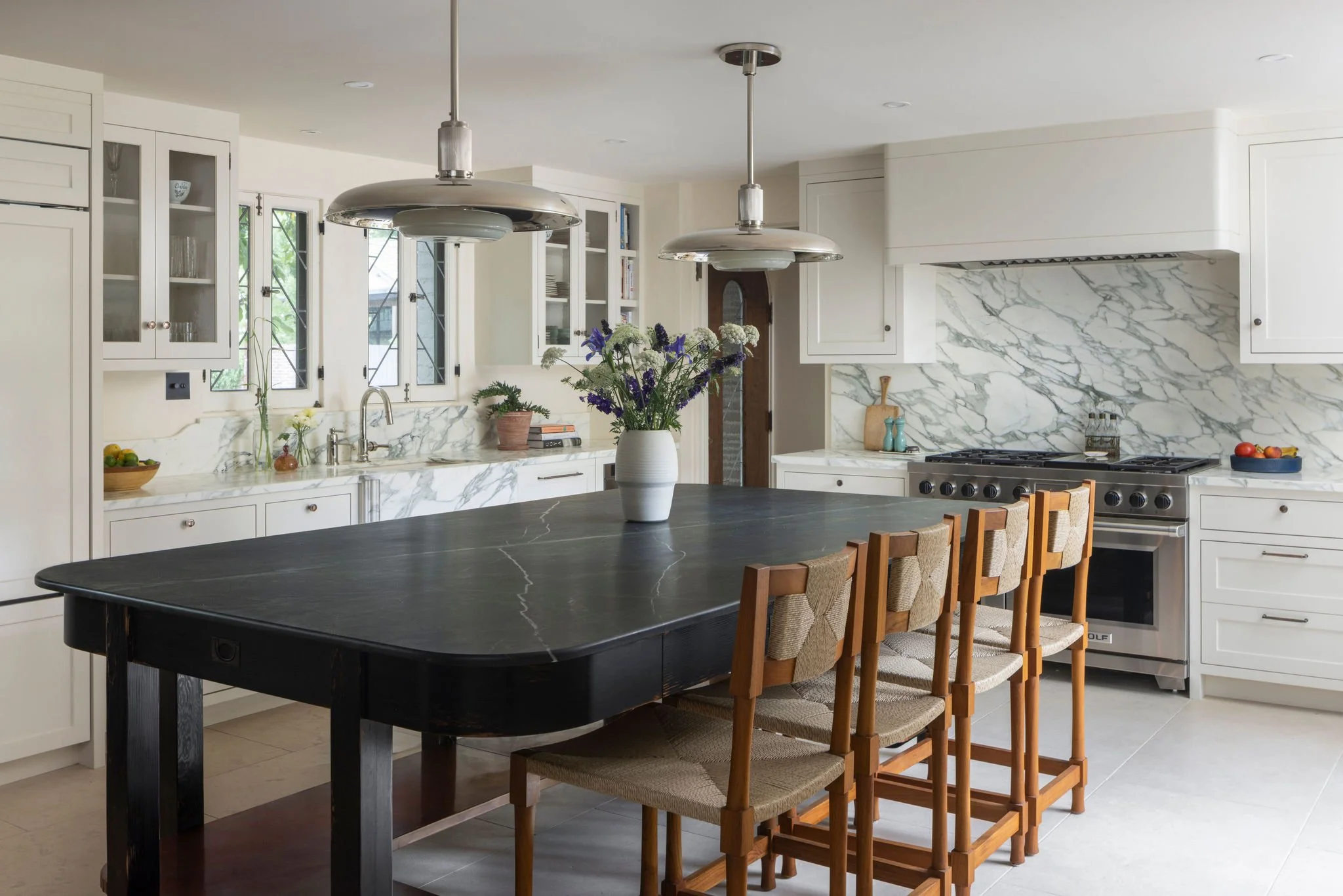 Modern kitchen with white cabinets, marble backsplash, black dining table with chairs, hanging pendant lights, and a vase of flowers on the table. Wolf range and Roman and Williams pendants. Custom barstools. Charleston Interior Designer. 