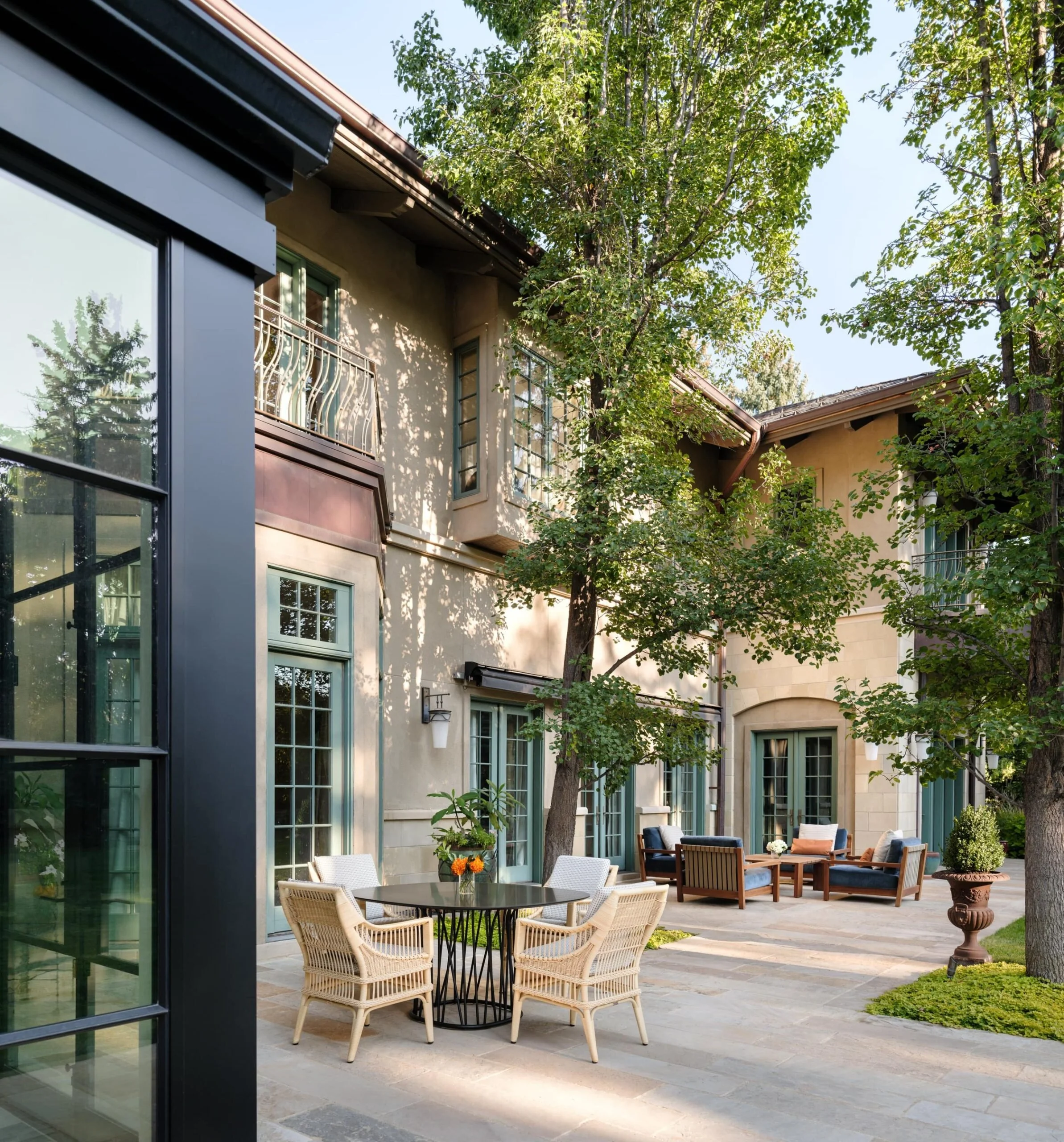 An outdoor patio of a beige multi-story house with large windows, surrounded by trees and greenery. The patio has a round table with chairs, and seating with blue cushions and a small table. Bonacina outdoor furniture. Charleston interior designer. 