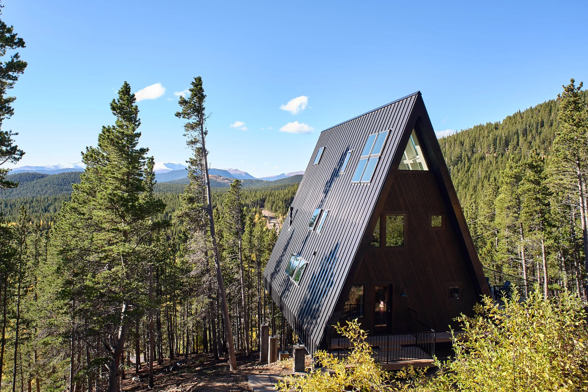 Unique A-frame house with large windows in a forested mountain landscape under a clear blue sky.