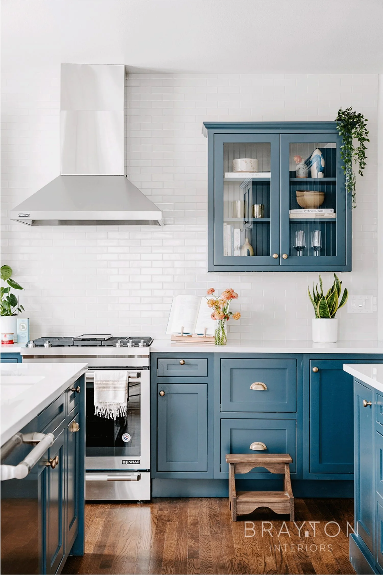 A modern kitchen with blue cabinetry, white countertops, a stainless steel stove and range hood, and decorative plants. Contains a glass-front wall cabinet, a vase of flowers, and a small wooden step stool on a wooden floor.
