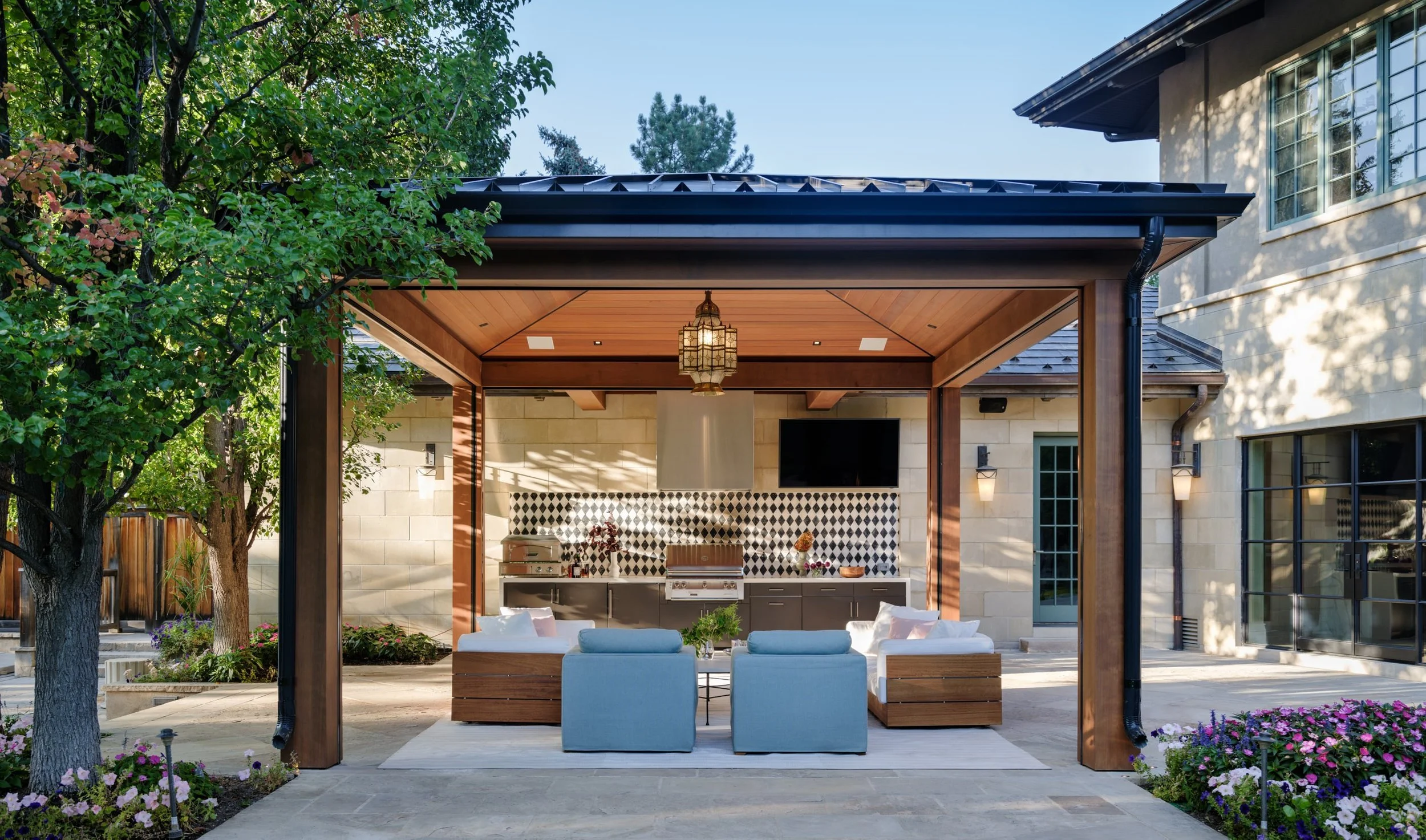 Outdoor patio with a built-in kitchen, seating area, trees, flower beds, and a house with big windows in the background. Bonacina outdoor furniture and custom pergola from Charleston Interior Designer. 