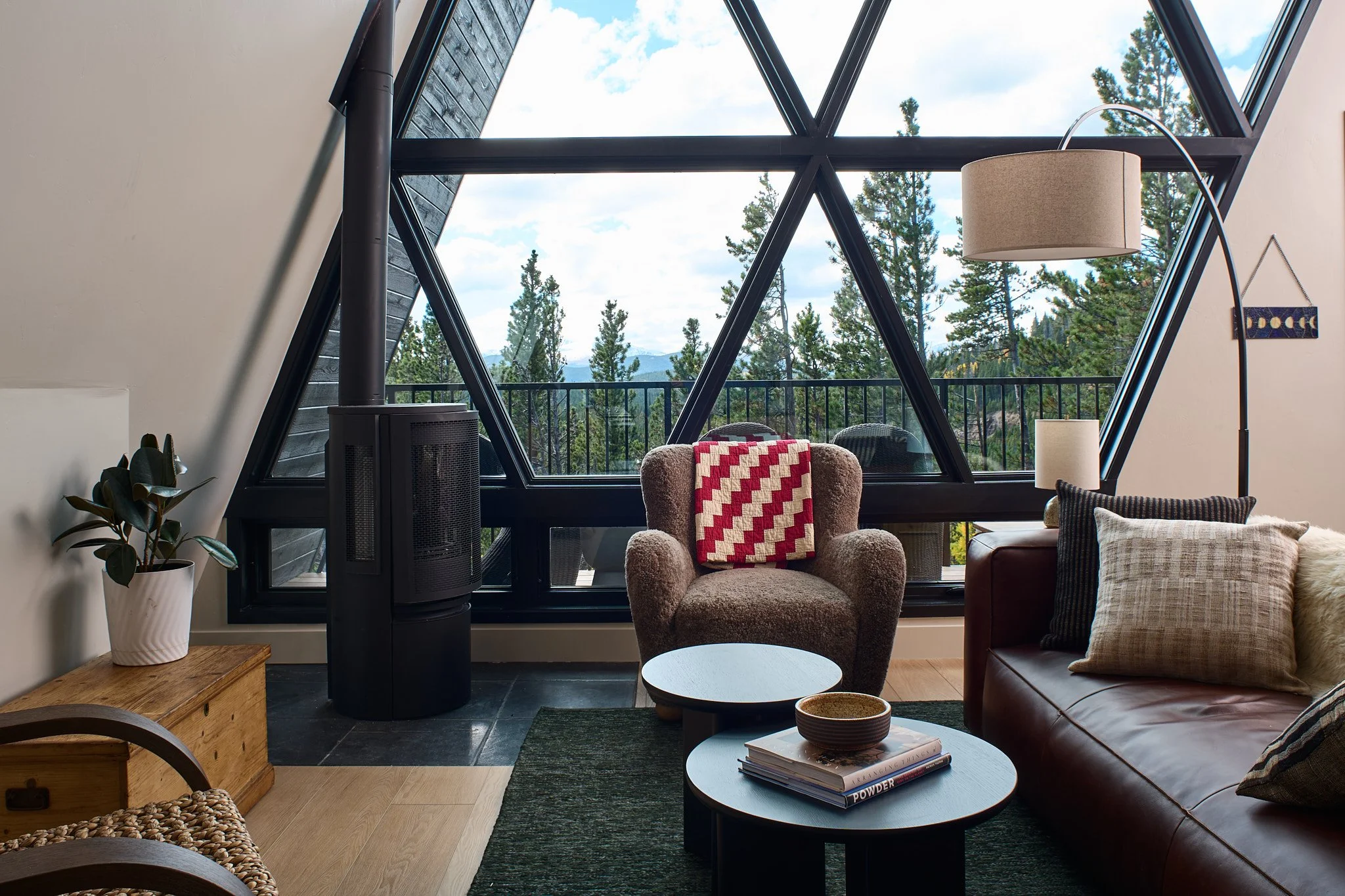 Living room with large triangular window, beige armchair with red checkered blanket, leather sofa with pillows, wooden side table with potted plant, lamps, and mountaintop view of trees outside.