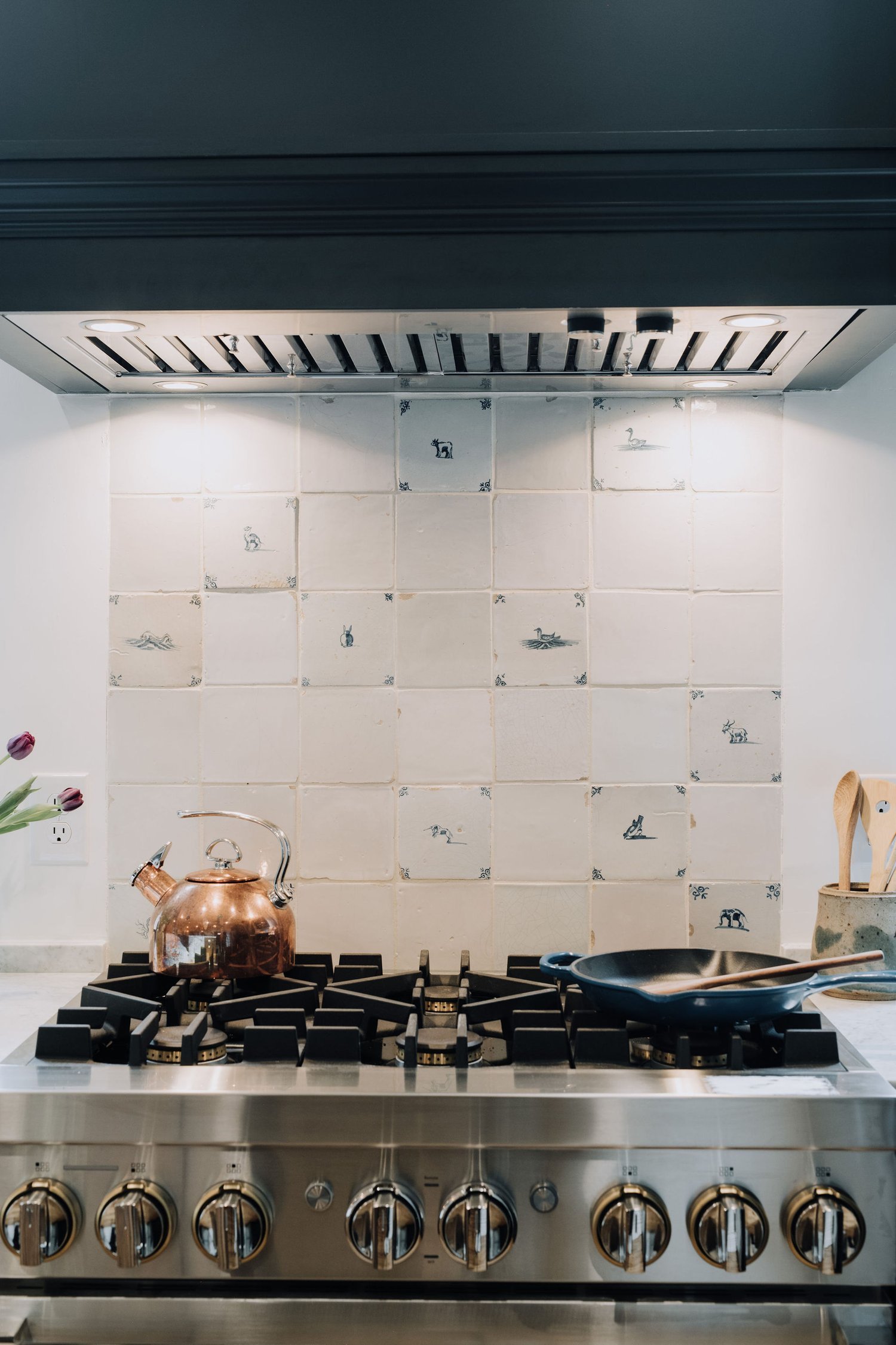 Stainless steel gas stove with copper kettle and frying pan, white tiled backsplash with blue and white wildlife illustrations, and a wooden utensil holder with spoons on the right. Charleston interior designer. 
