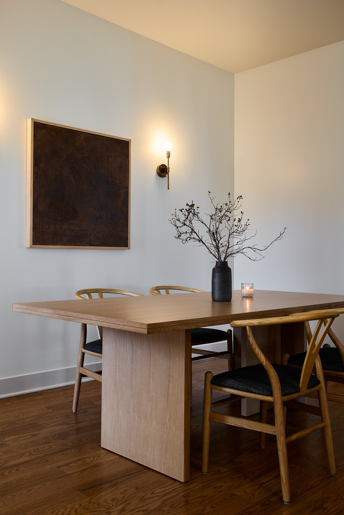 A minimalist dining room with a wooden table, three wooden chairs with black cushions, a black vase with branches, a candle, and a wall-mounted light fixture.