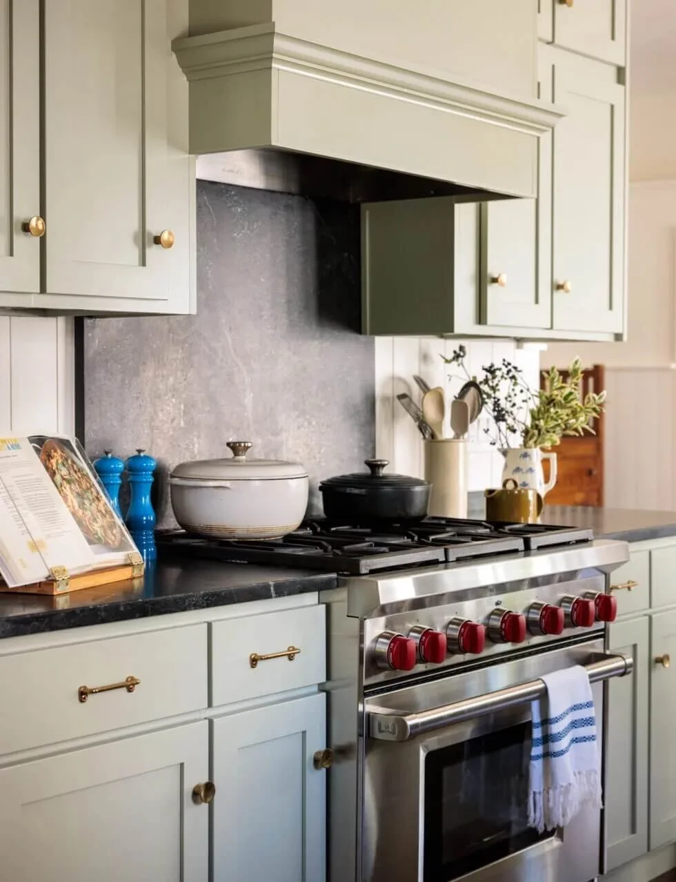 Kitchen with light green cabinets, black countertop, gray backsplash, and a stainless steel stove with red knobs. Items include a cookbook, blue spice jars, white and black pots, and a vase with greenery.