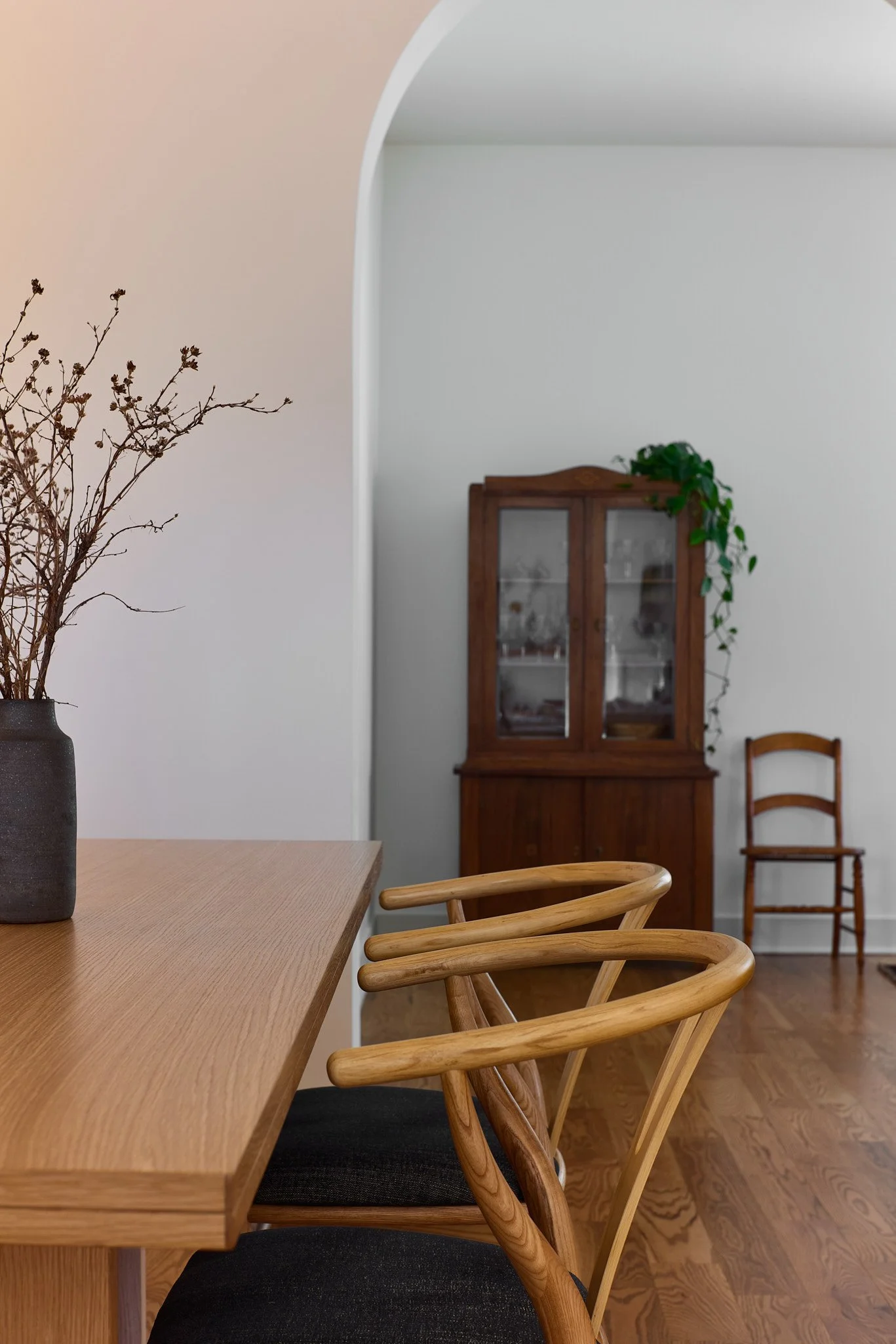 A wooden dining table with black cushioned chairs, a vase with dried branches, a wooden cabinet with glass doors, a wooden chair, and a green potted plant on top, set in a room with hardwood floors and white walls.
