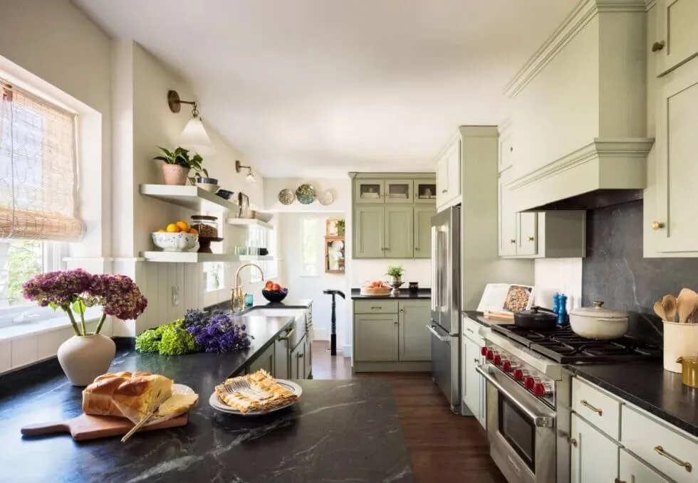 Bright kitchen with light green cabinets, black countertops, a window with a woven shade, and shelves with decorative items and fruit. On the counter, there is a vase of pink flowers, a loaf of bread, and a cheese platter. Modern appliances include a stainless steel refrigerator and a gas stove. The room has a warm and inviting atmosphere with natural light.