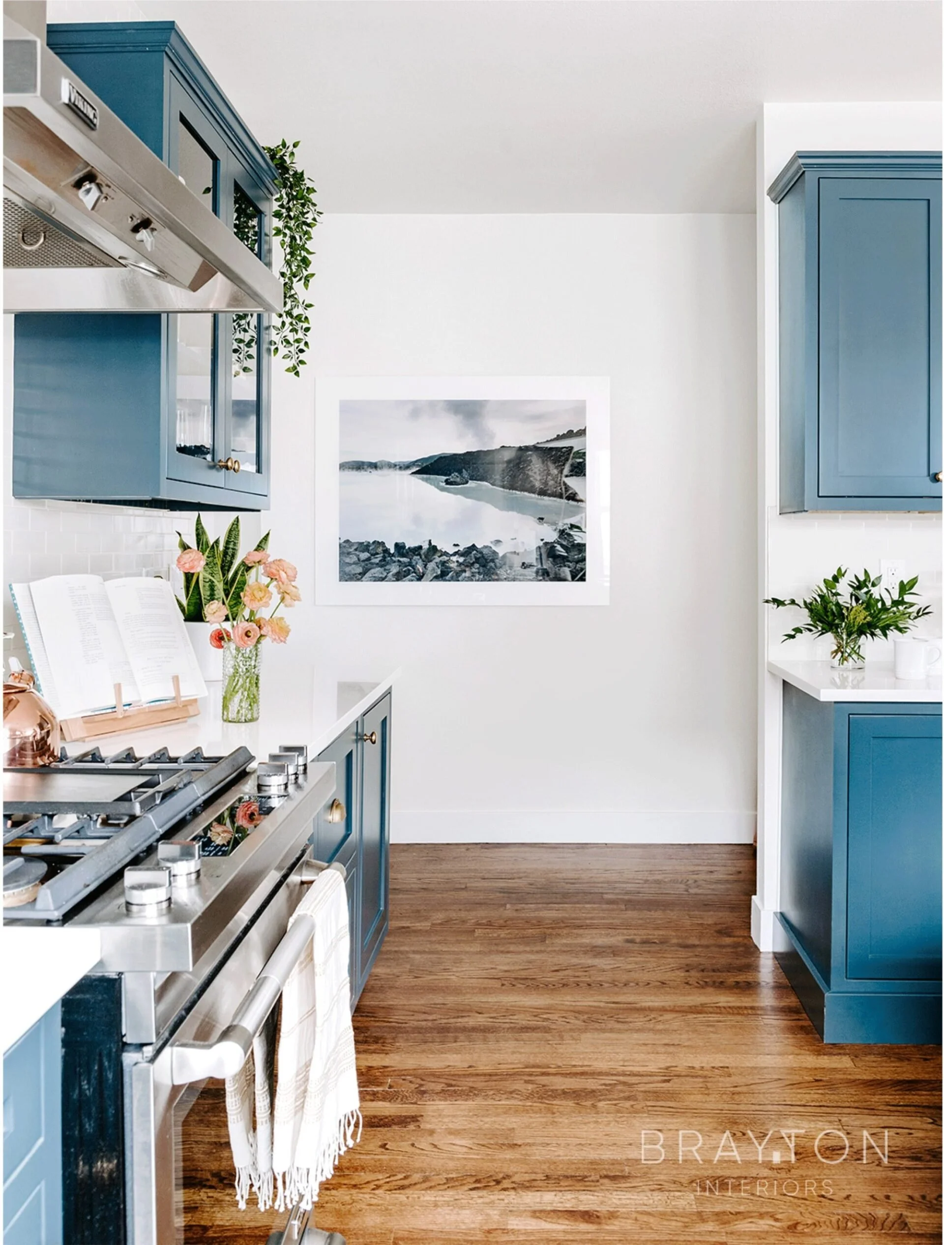 Kitchen with blue cabinets, stainless steel stove, white countertops, wood flooring, pink flowers in a glass vase, wall art of a lake and mountains, and green plants. Home renovation by Charleston interior designer. 