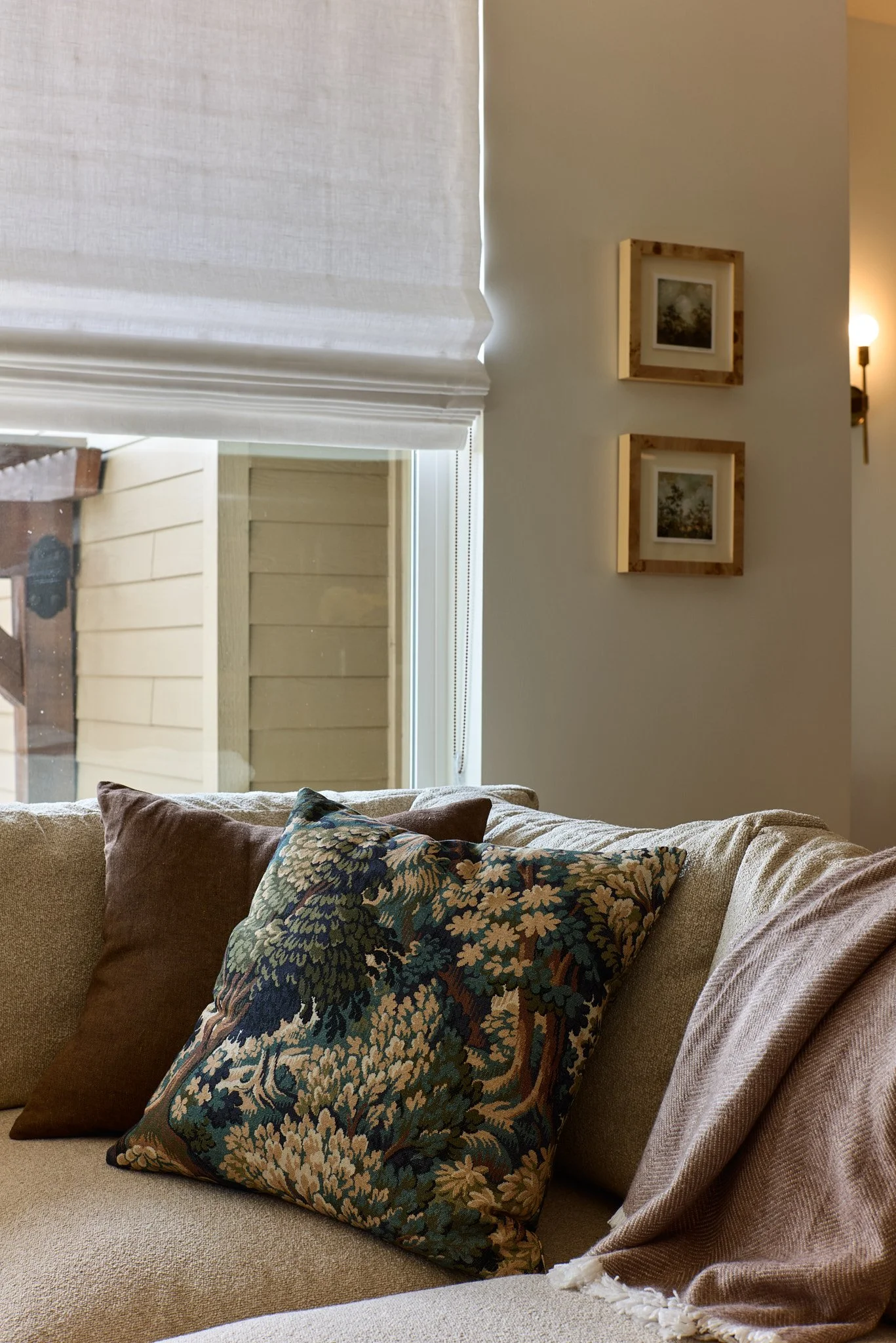 A cozy living room corner with a beige sofa, decorative pillows, a window with cut blinds, and framed artwork on the wall.