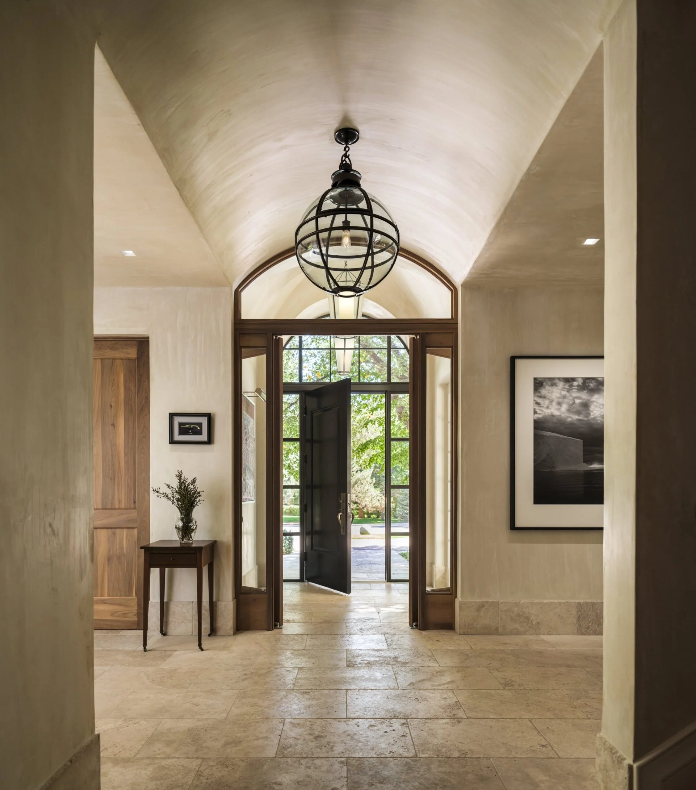 Elegant foyer with arched ceiling, hanging light fixture, framed black and white photograph, small wooden table with a vase and greenery, and glass-paneled front door leading outside to a sunny garden. Charleston interior designer Sarah Hart.
