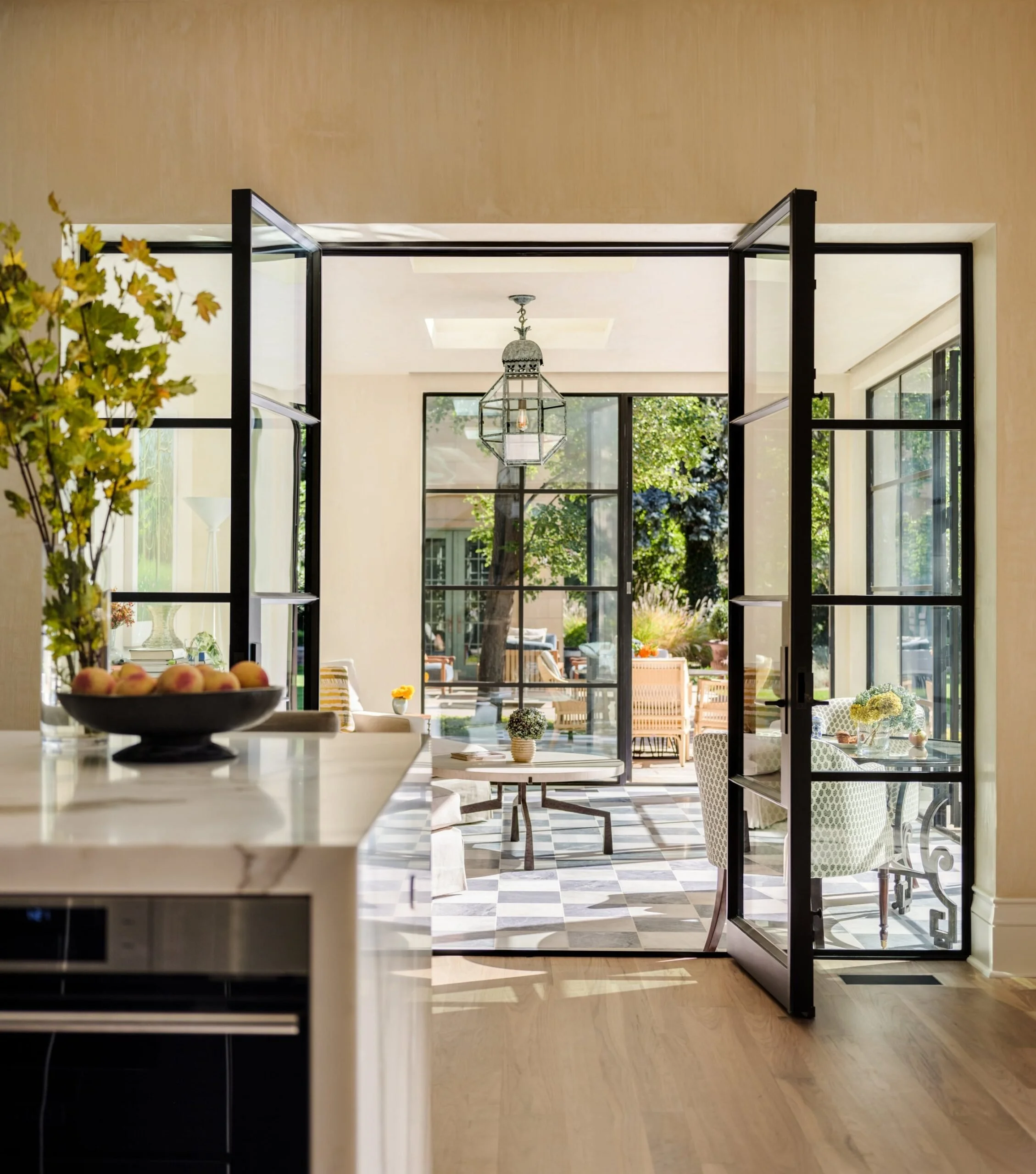 Open black-framed glass doors lead to a sunlit patio with outdoor furniture and greenery. Charleston interior designer Sarah Hart Interior Design. 