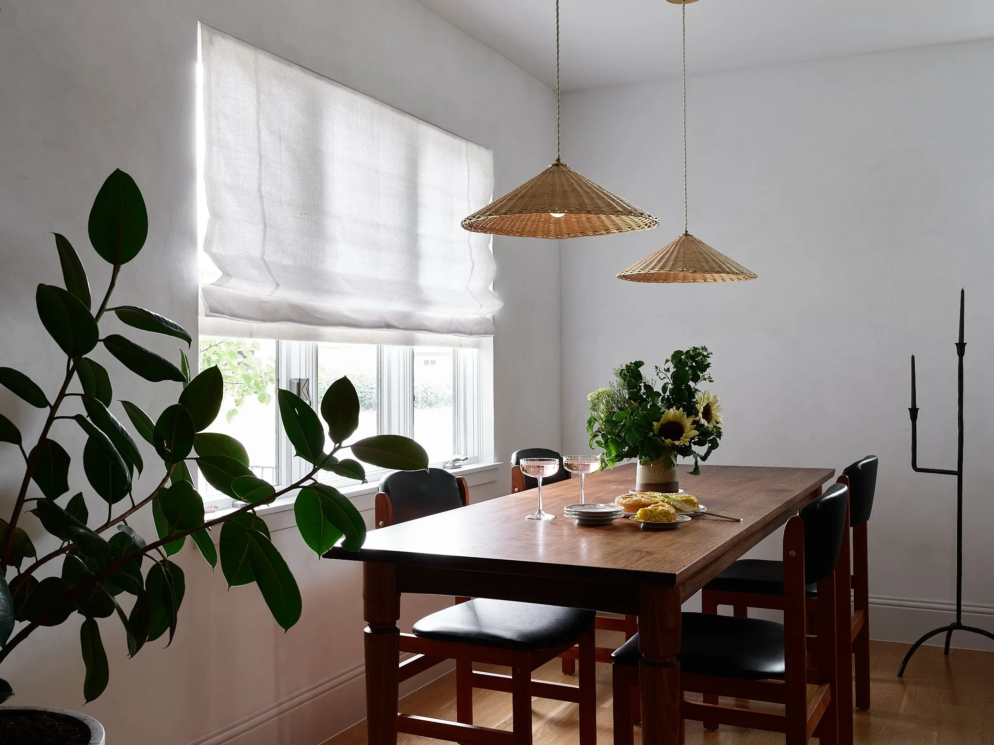 A dining room with a wooden table set with a bouquet of sunflowers, empty wine glasses, and some plates and food. There are four black chairs around the table. A large window with a white Roman shade is on the left, and two wicker pendant lights hang from the ceiling. There is a large leafy houseplant on the left and a black floor candle holder on the right.