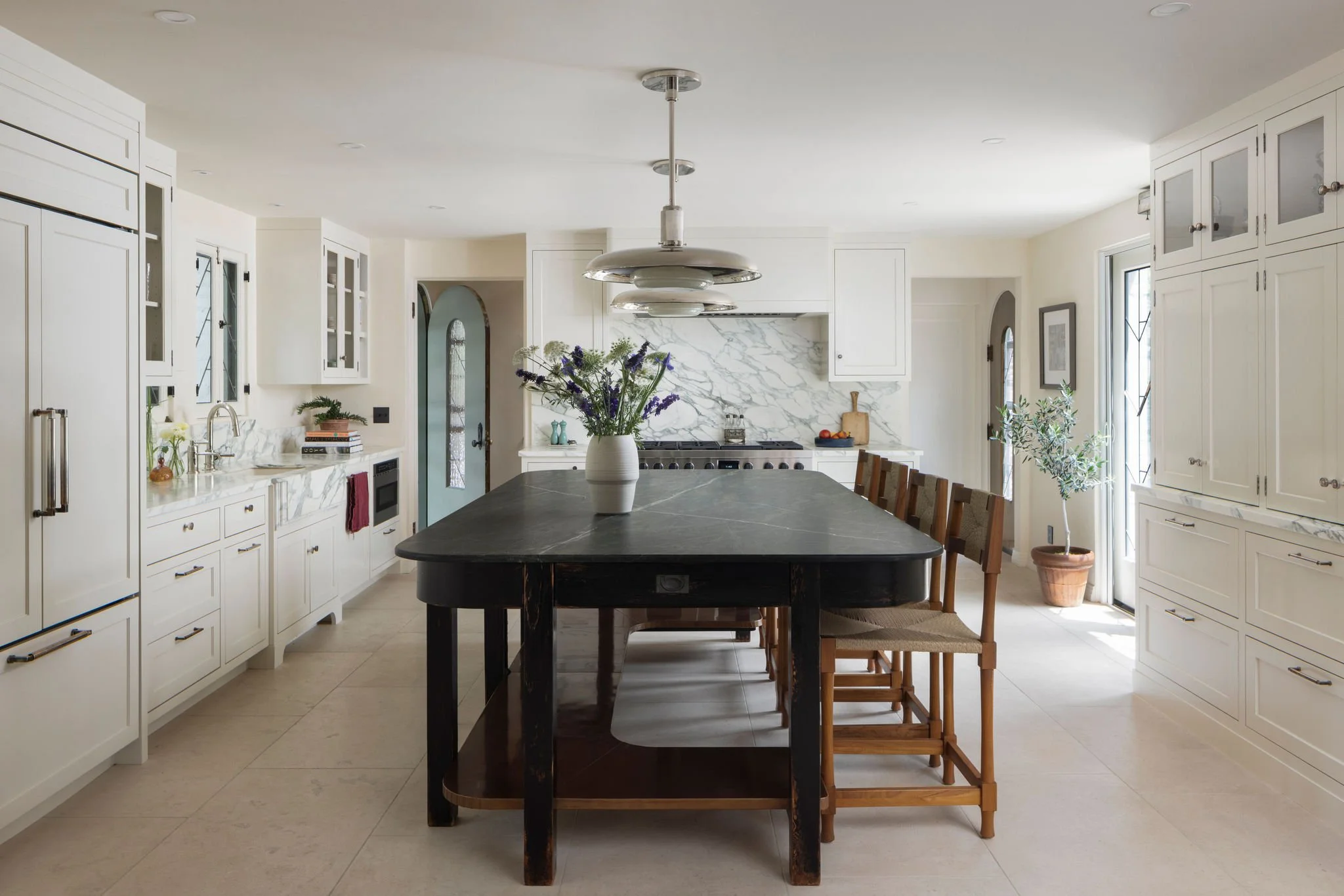 Bright kitchen with white cabinets, a black kitchen island with a vase of flowers, a marble backsplash, and natural light from windows and a glass door. Calacatta turquoise marble backsplash and Roman and Williams pendants. Charleston Interior Design