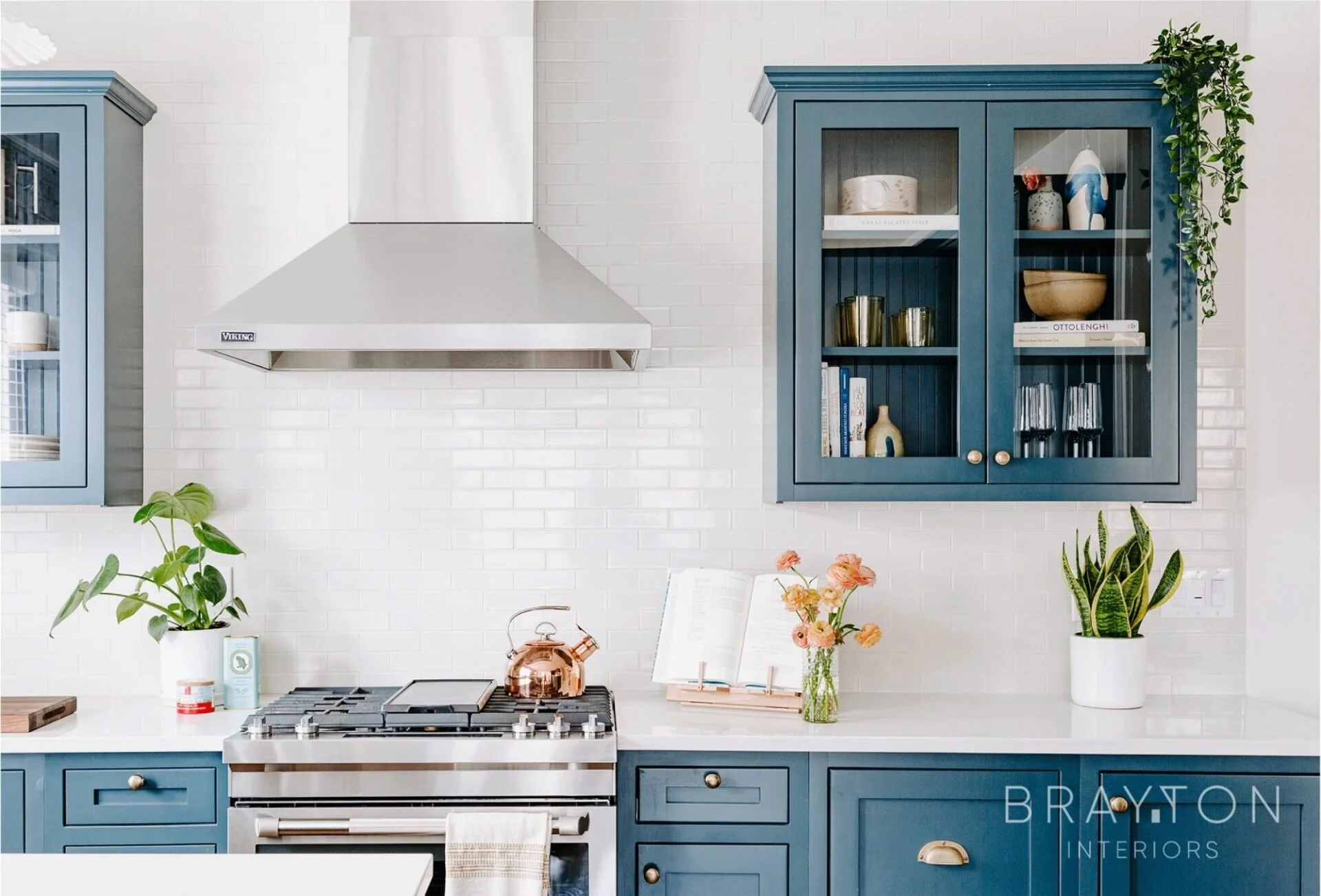 A modern kitchen with blue cabinets, white countertop, stainless steel stove, copper kettle, open cookbook, glass vase with orange flowers, and two potted green plants on white tile backsplash. Custom kitchen by Charleston designer. 