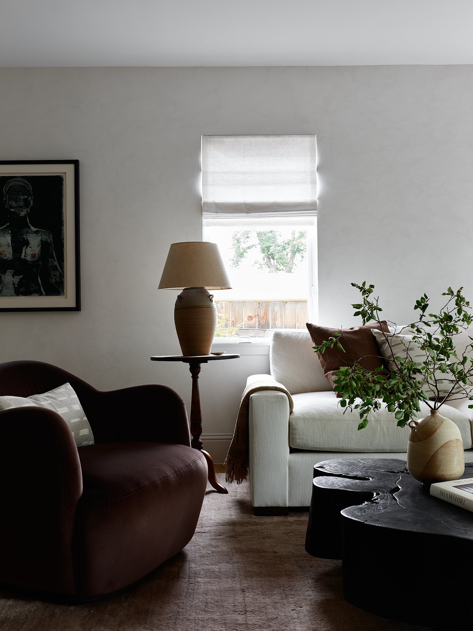 Living room with a white sofa, dark armchair, side table with a lamp, a window with blinds, a framed artwork on the wall, and decorative plants on a coffee table.