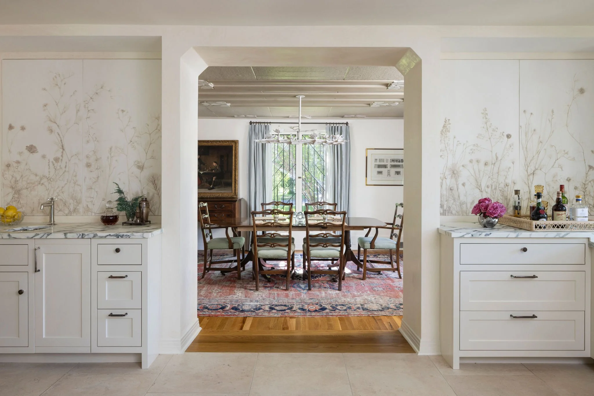 View of a dining room with a large wooden table and eight chairs, seen through an open archway from a kitchen area with white cabinets and marble counters, decorated with flowers, bottles, and lemons. Charleston interior designer. 