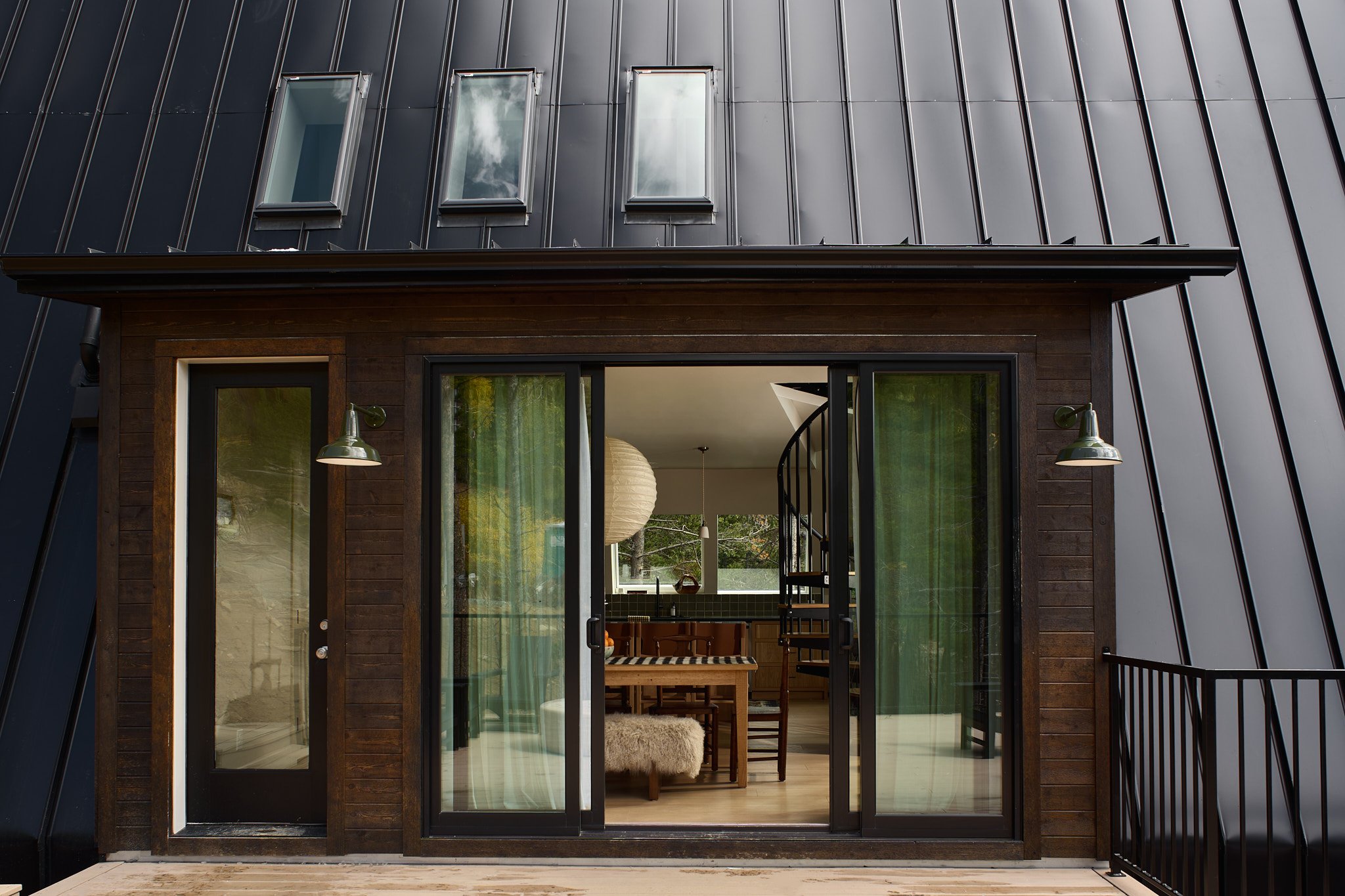 The image shows a modern house exterior with dark-colored metal siding, three small windows on the top, and a large sliding glass door leading into the interior. Inside, there is a dining table with chairs, a spiral staircase, and a large paper lantern hanging from the ceiling.