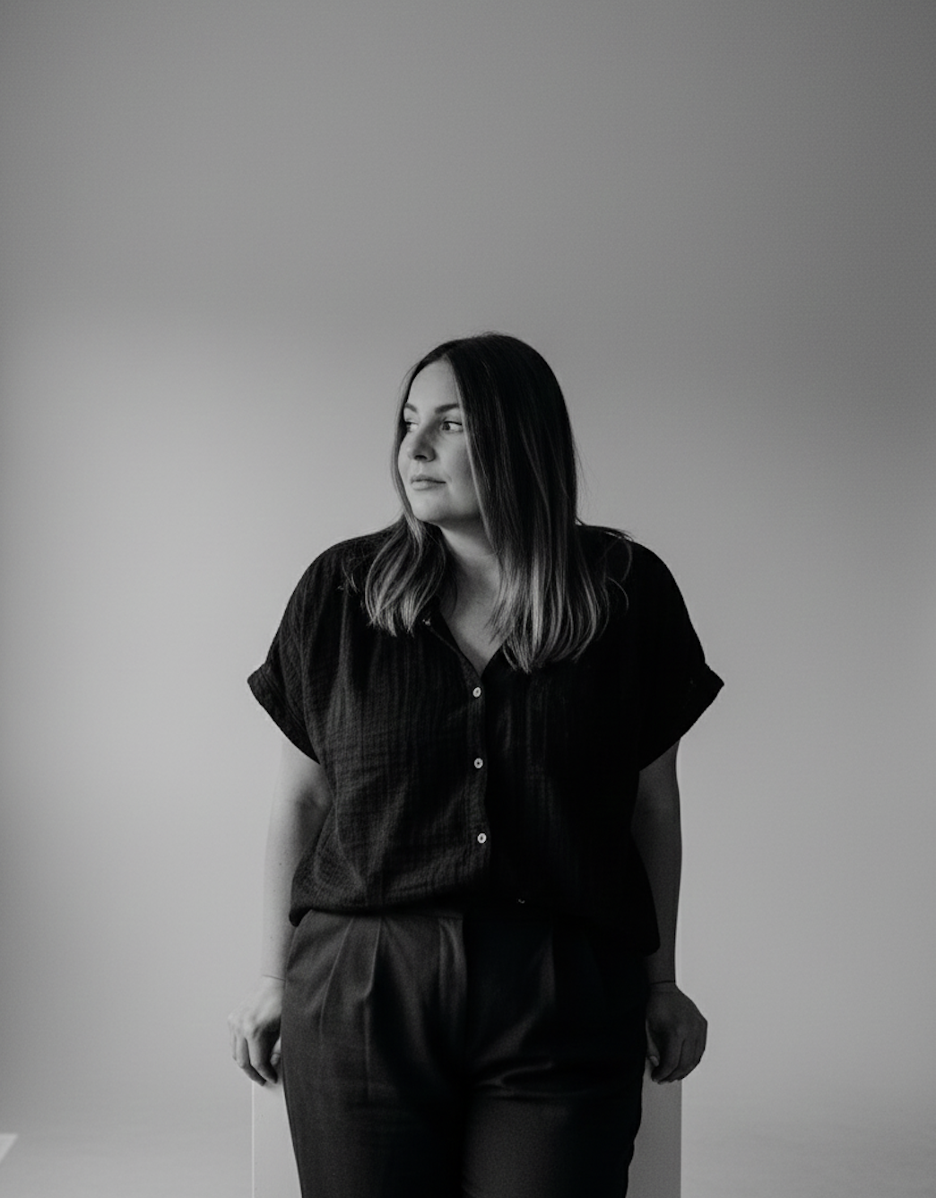 Black and white photo of a woman with shoulder-length hair wearing a dark, short-sleeved blouse and dark pants, standing against a plain background.