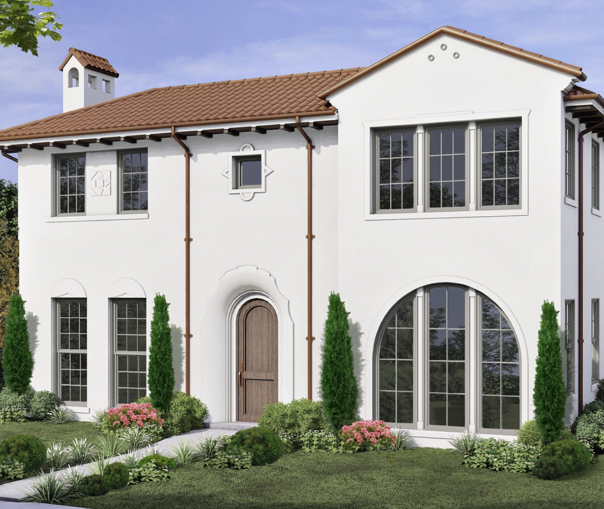 A two-story white house with brown tiled roof, arched and rectangular windows, a wooden front door, and landscaped greenery with shrubs and flowers in front.