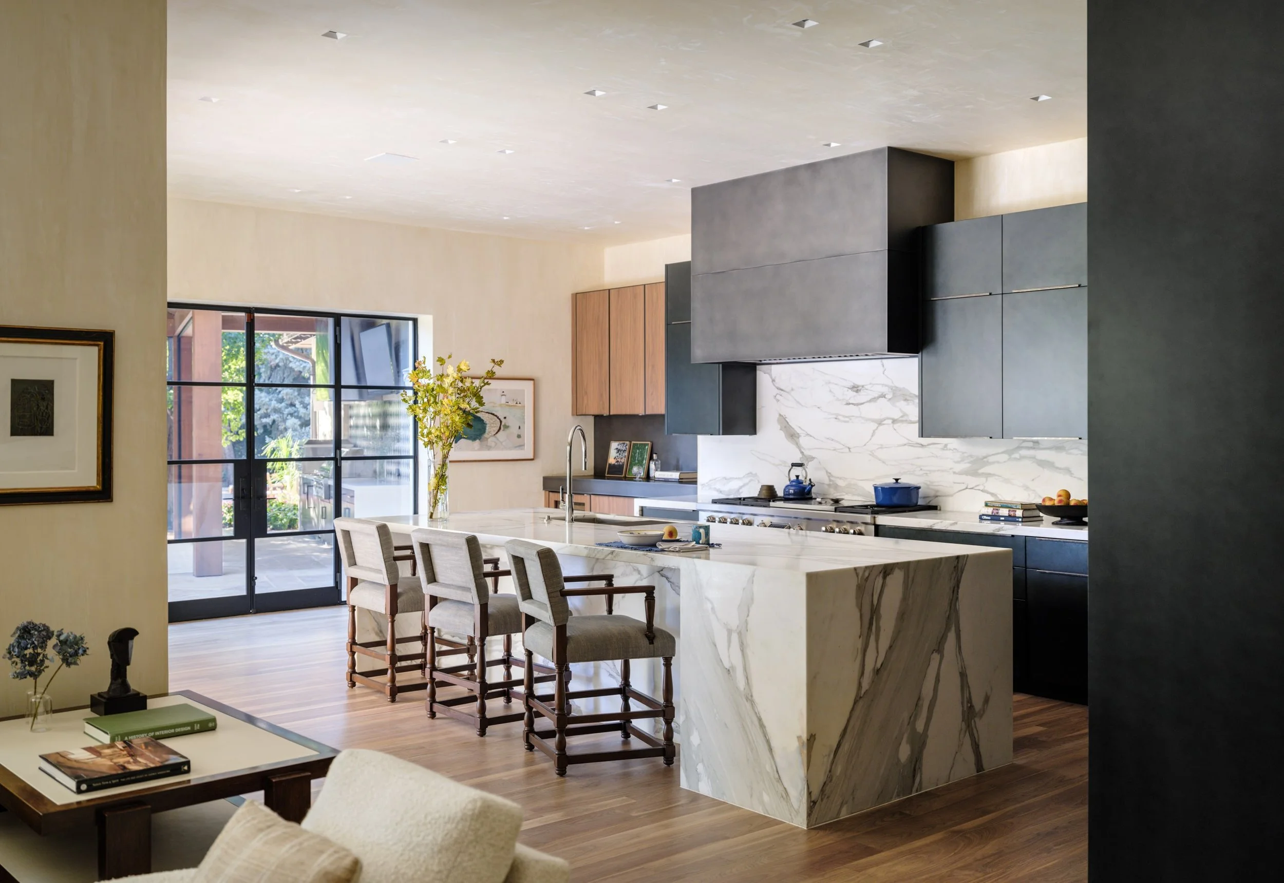 Modern open-concept kitchen with white marble island and backsplash, dark gray cabinet upper units, wooden lower cabinets, a blue kettle and pots, bar stools, and sliding glass doors leading outside.