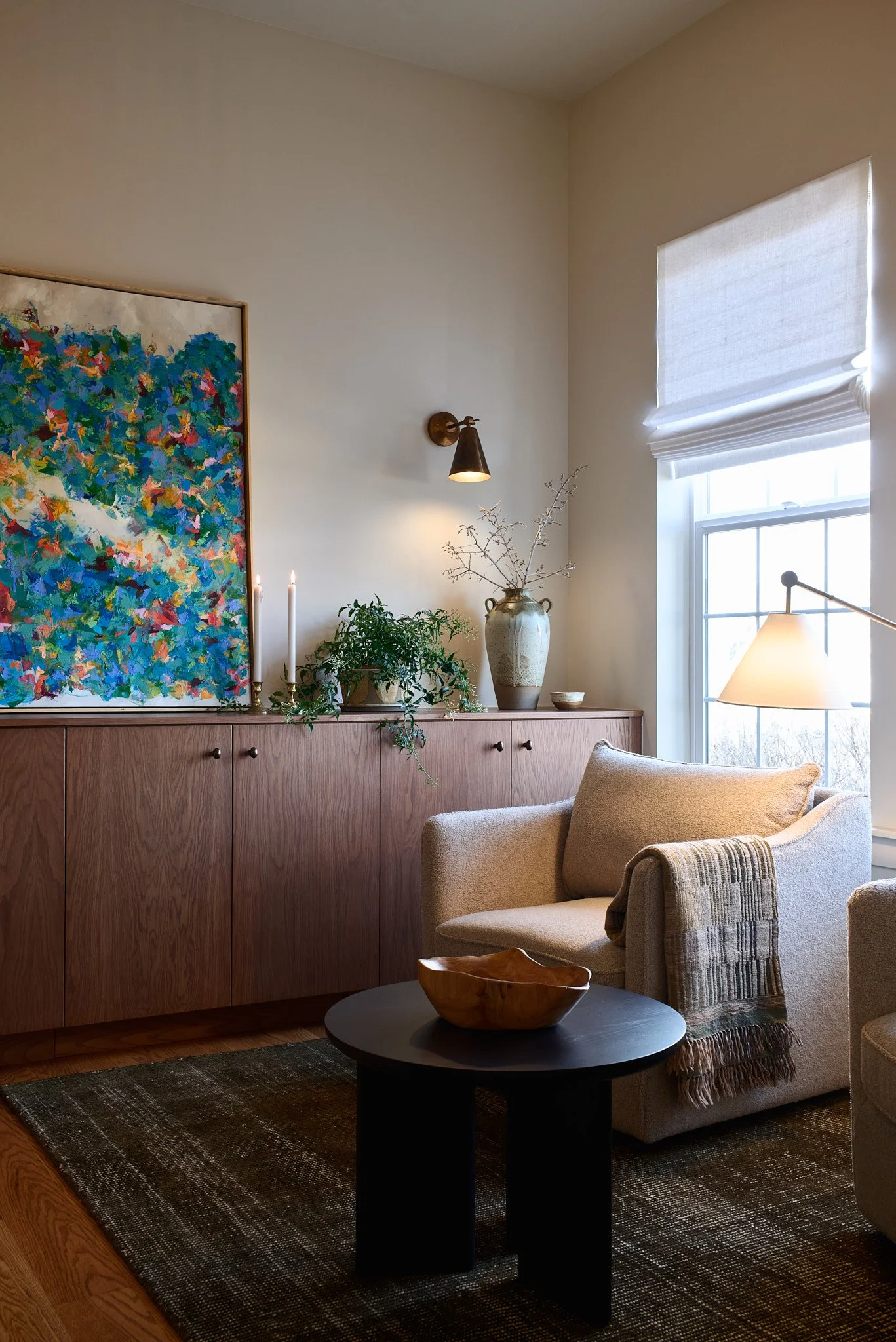 Living room with beige armchair, black coffee table, wooden cabinet with plants and artwork, window with white shades, beige wall with wall lamp, and area rug.