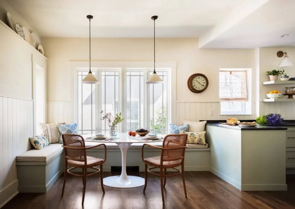 Bright kitchen and dining area with large windows, a round dining table with two chairs, built-in bench seating with pillows, and shelves with dishes and plants. Historic renovation by Charleston Interior Designer.