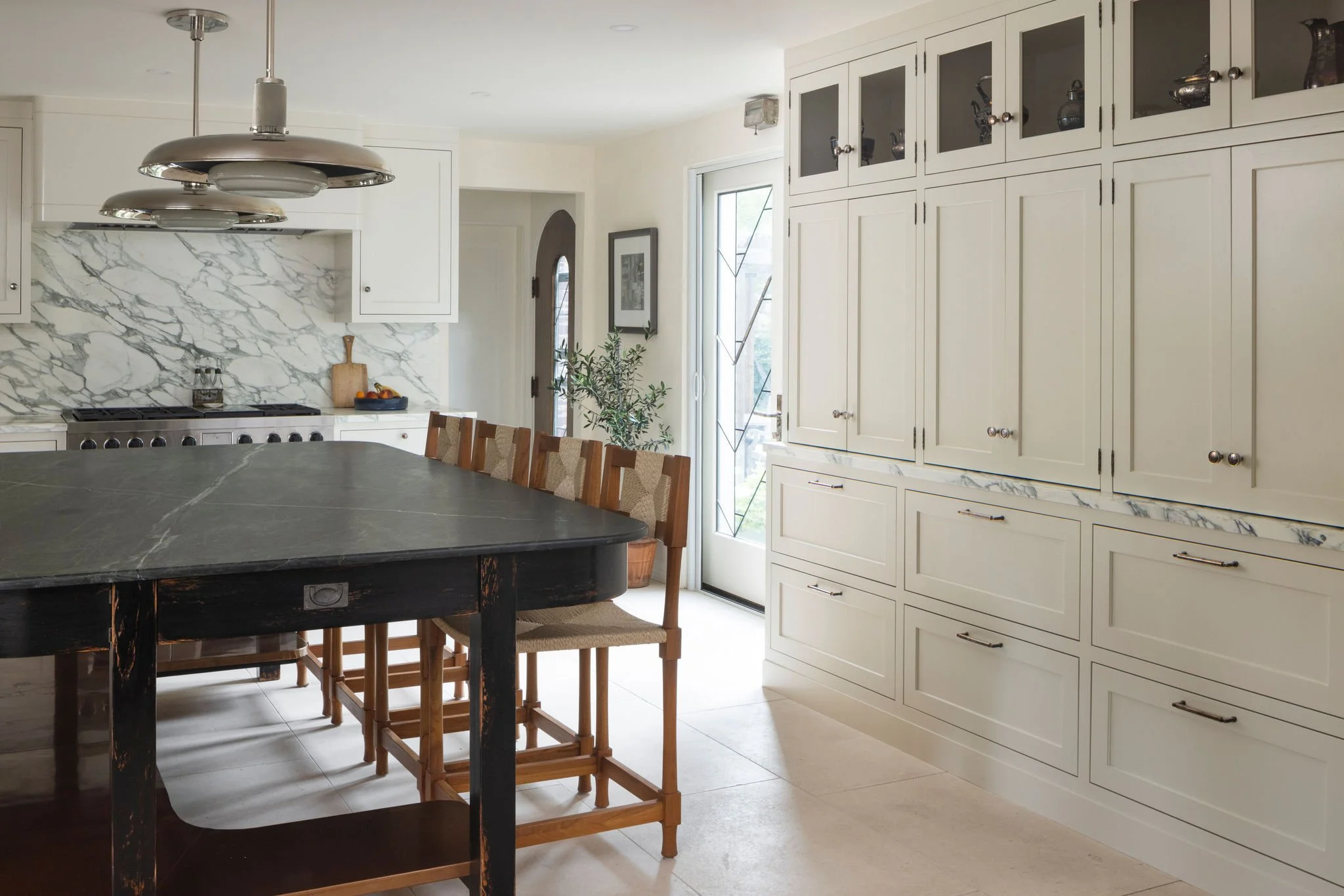 Modern kitchen with white cabinets, marble backsplash, black island with a marble top, and wooden chairs. Roman and Williams pendants and calacatta turquoise marble countertops. 
