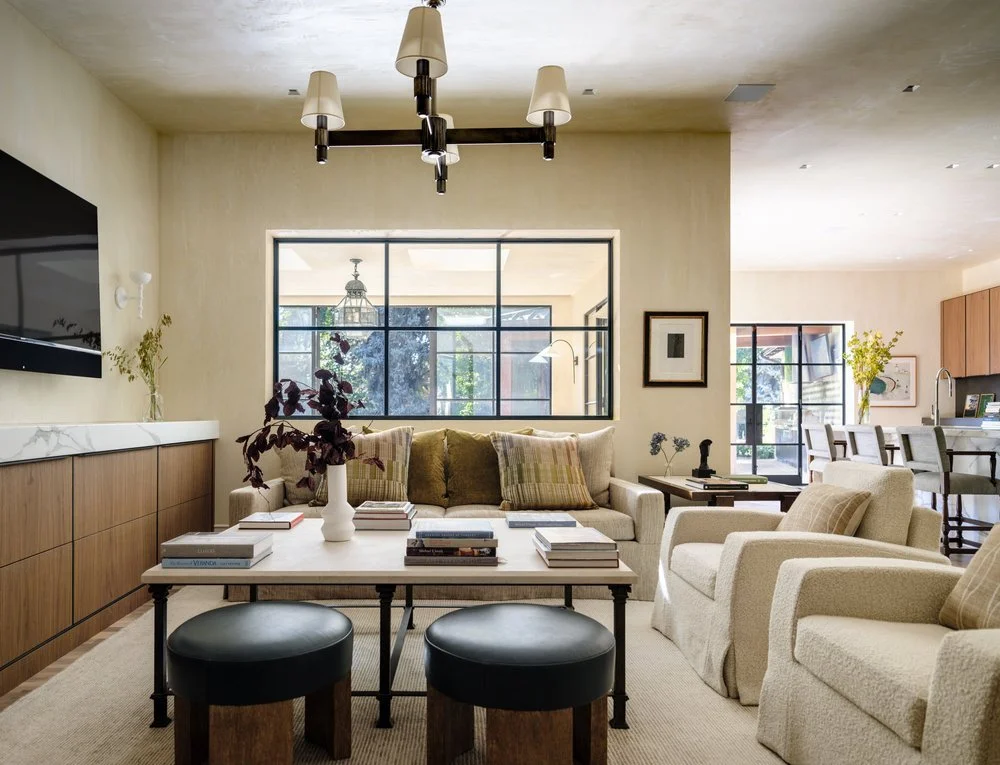 Living room with beige armchairs, a beige sofa, a white marble-topped sideboard, a white coffee table with books and a vase, and a wall-mounted TV. Large windows and an open kitchen in the background. Charleston interior designer Sarah Hart.