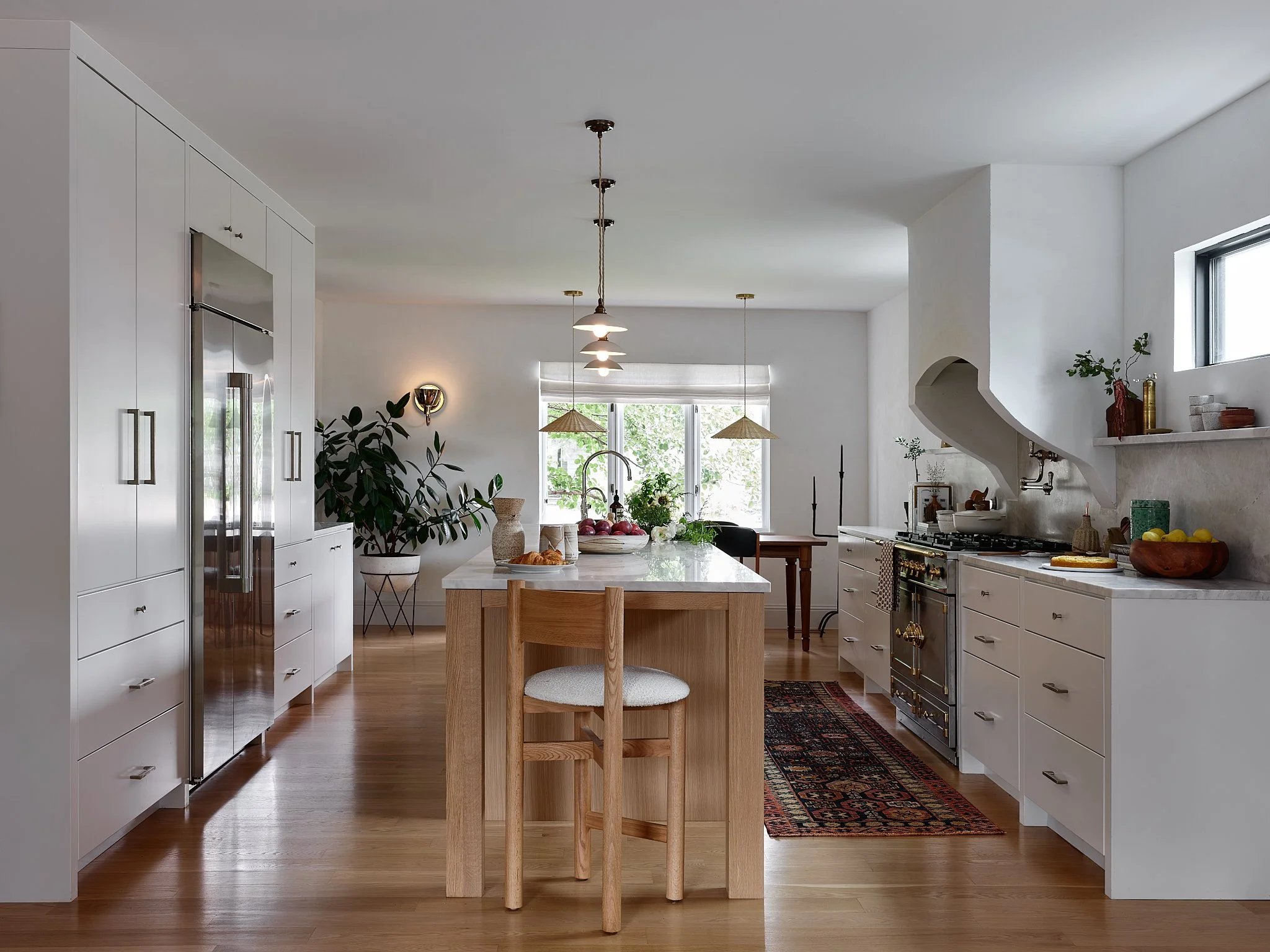 Modern kitchen with white cabinets, a wooden island, and hardwood floors. Decor includes plants, bowls of fruit, and pendant lighting.
