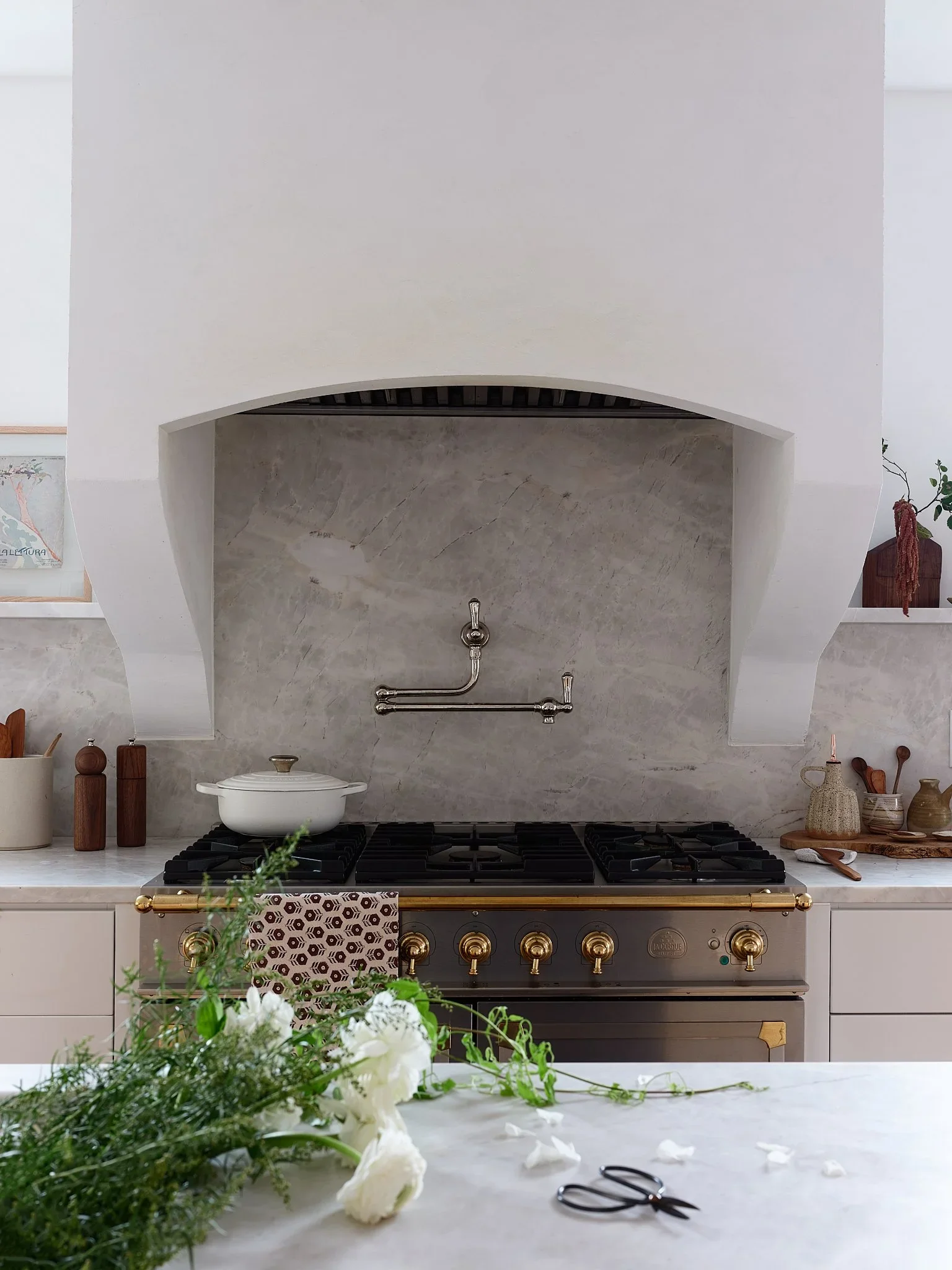 Kitchen with a marble backsplash and a large stovetop. White and cream-colored decor with plants, jars, and a pair of scissors on a marble counter in the foreground.